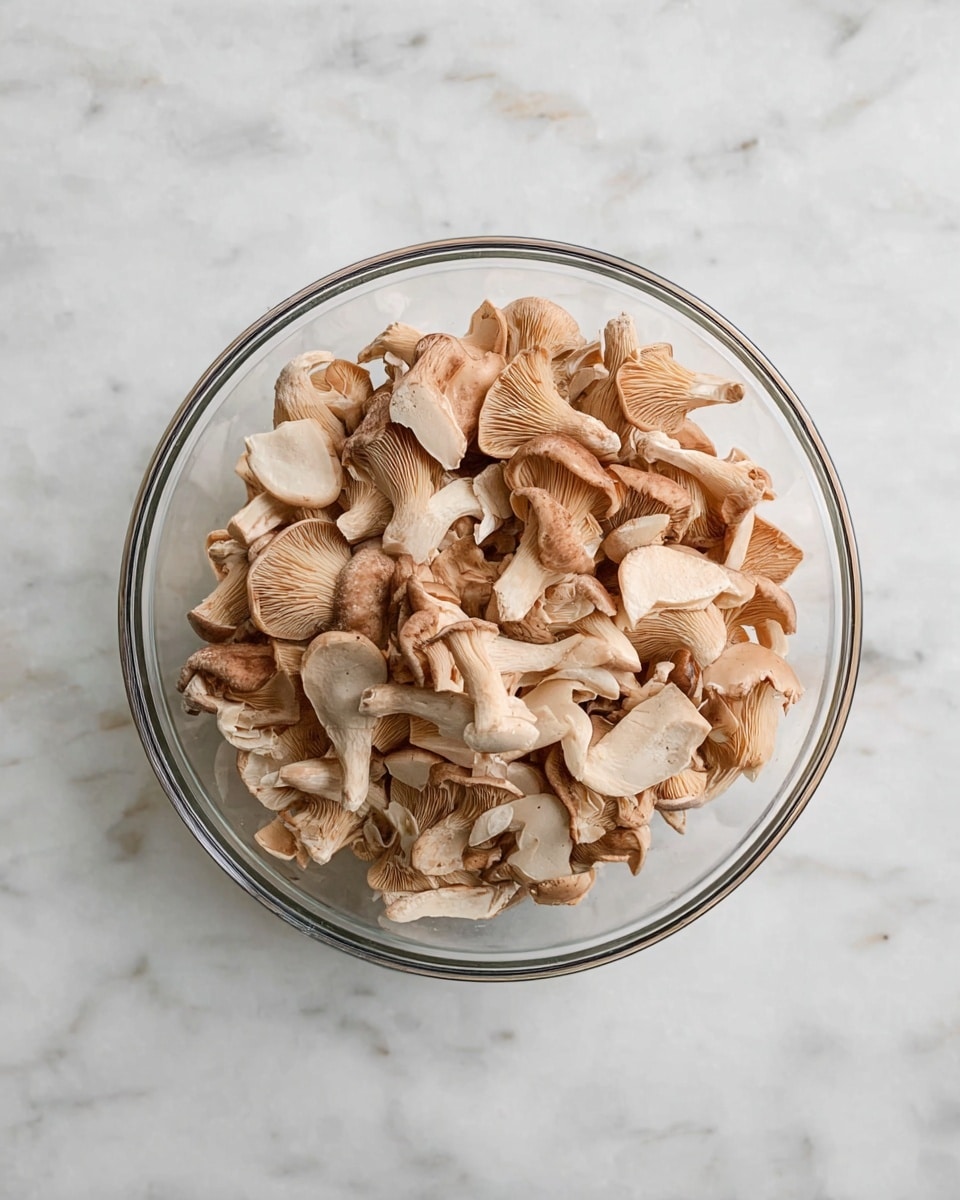 A clear glass bowl filled with a mix of raw mushrooms in various shapes and sizes, showing a range of light browns, beige, and creamy off-white colors, arranged in a slightly uneven pile that fills the bowl almost to the top, placed on a white marbled surface photo taken with an iphone --ar 4:5 --v 7