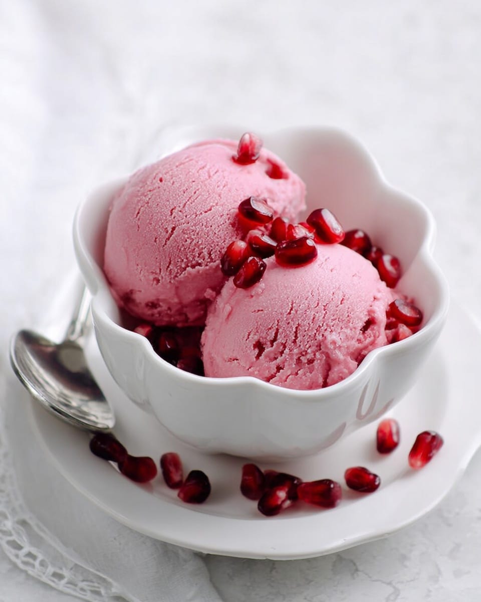 Two scoops of pink ice cream with a smooth, slightly rough texture are placed in a white bowl with wavy edges. The ice cream is garnished with bright red pomegranate seeds that add small bursts of color on and around the scoops. The bowl sits on a white plate that carries some scattered pomegranate seeds and a shiny silver spoon on the left side. The background is a white marbled surface that gives a clean and soft look. Photo taken with an iphone --ar 4:5 --v 7