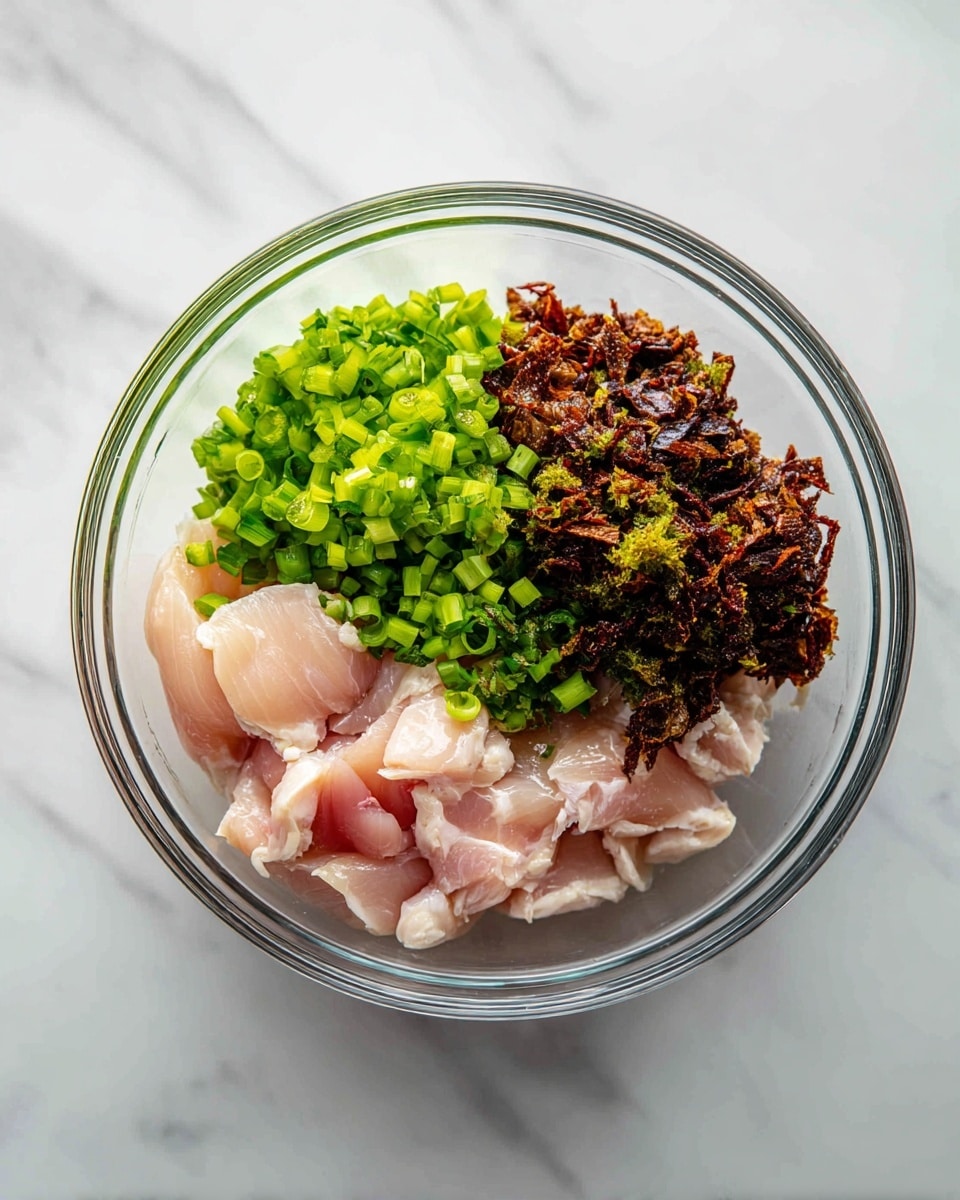 A clear glass bowl sits on a white marbled surface, filled with three main layers: thin slices of light pink raw chicken pieces forming the base, a layer of finely chopped bright green spring onions placed on one side, and a pile of dark brown crispy bits, likely fried garlic or onions, arranged beside the green onions. The ingredients are not mixed but kept separate in the bowl, showing the contrast between the soft pale chicken, the fresh green onions, and the crunchy dark brown topping. Photo taken with an iphone --ar 4:5 --v 7