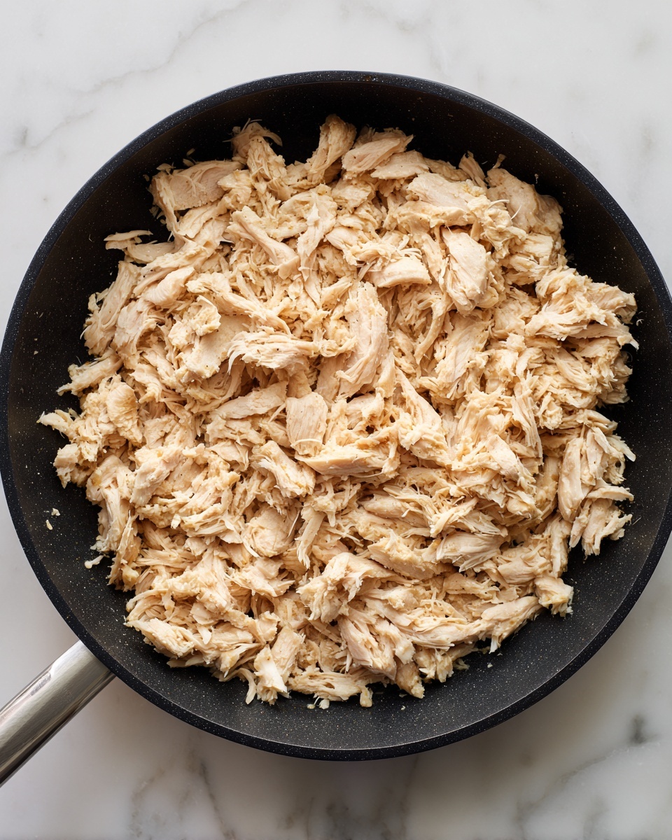 The image shows a black frying pan filled with a single layer of shredded cooked chicken that is light brown and white in color, giving a rough and crumbly texture. The chicken pieces are spread evenly across the pan's surface, covering it fully. The pan sits on a white marbled surface. Photo taken with an iphone --ar 4:5 --v 7