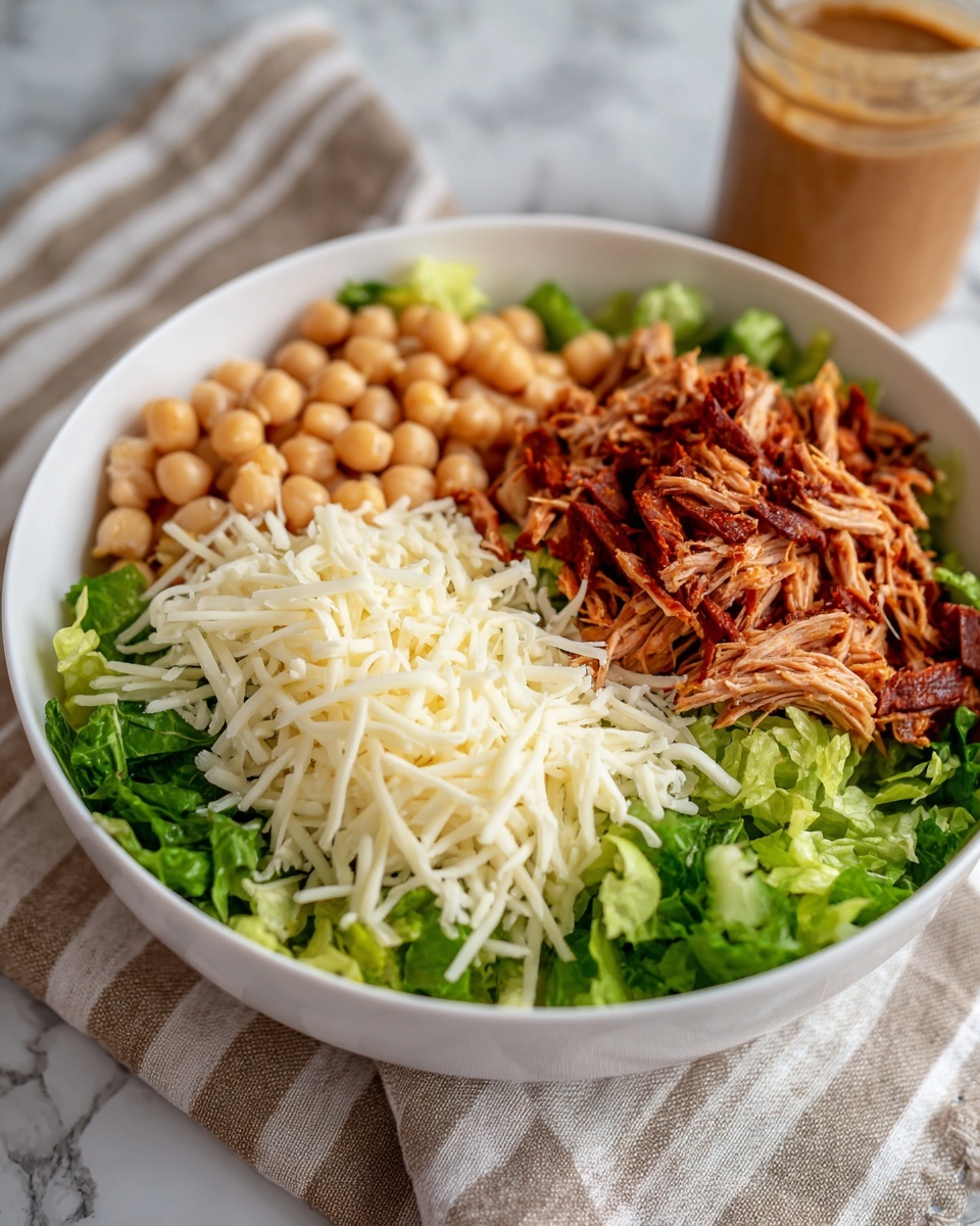 A white bowl with four clear layers is placed on a white marbled surface with a striped beige and grey cloth beneath it. The bottom layer is fresh chopped green lettuce covering a third of the bowl to the right side. Next to the lettuce layer, there are round beige chickpeas filling about a sixth of the bowl. To the left of the chickpeas, a neat pile of white shredded cheese takes up another sixth of the bowl. On top of the lettuce, chickpeas, and cheese, there is a generous layer of thin, dark red strips of meat that cover these areas. In the background, there is a jar with a thick, light brown dressing. The photo taken with an iphone --ar 4:5 --v 7