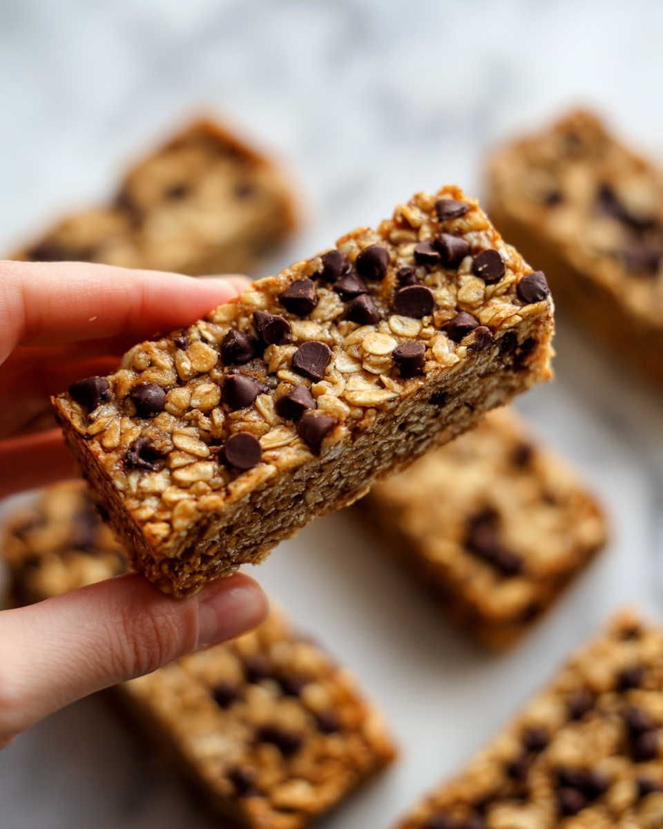 A close-up view of a rectangular granola bar with a golden-brown color, filled with visible oats and dark chocolate chips spread evenly on top and inside the bar, giving it a chewy texture. The granola bar is being held gently by a woman's hand, showcasing the thickness and firmness of the bar. Multiple similar bars are blurred in the white marbled background, emphasizing the focus on the granola bar held in the front. Photo taken with an iphone --ar 4:5 --v 7