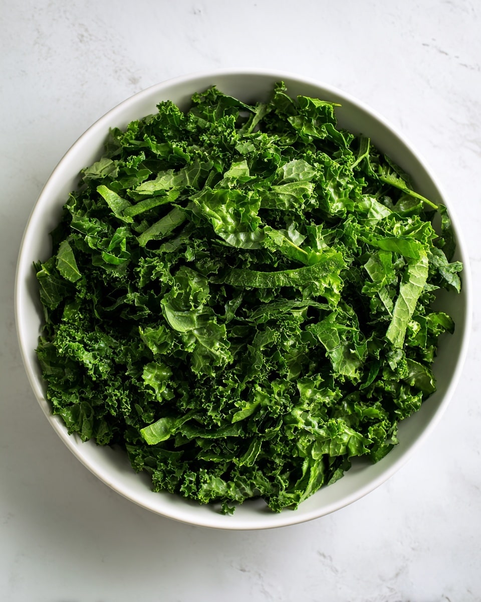 A large white bowl filled with many thin strips of dark green leafy kale. The kale is evenly shredded and layered loosely inside the bowl, showing a textured and slightly curly surface. The bowl sits on a white marbled surface, giving a clean and bright background to the fresh kale. photo taken with an iphone --ar 4:5 --v 7