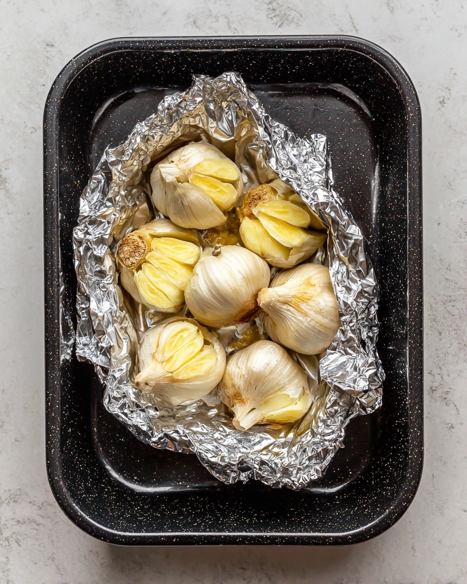 A black speckled roasting pan sits on a white marbled surface, holding a small bundle of garlic heads wrapped partially in shiny silver aluminum foil. The garlic heads are cut across the top, revealing a soft light golden-yellow color of the cloves inside, some of which have a smooth, slightly caramelized texture. The foil is crinkled around the garlic, catching light and adding brightness to the dark pan below. Photo taken with an iphone --ar 4:5 --v 7