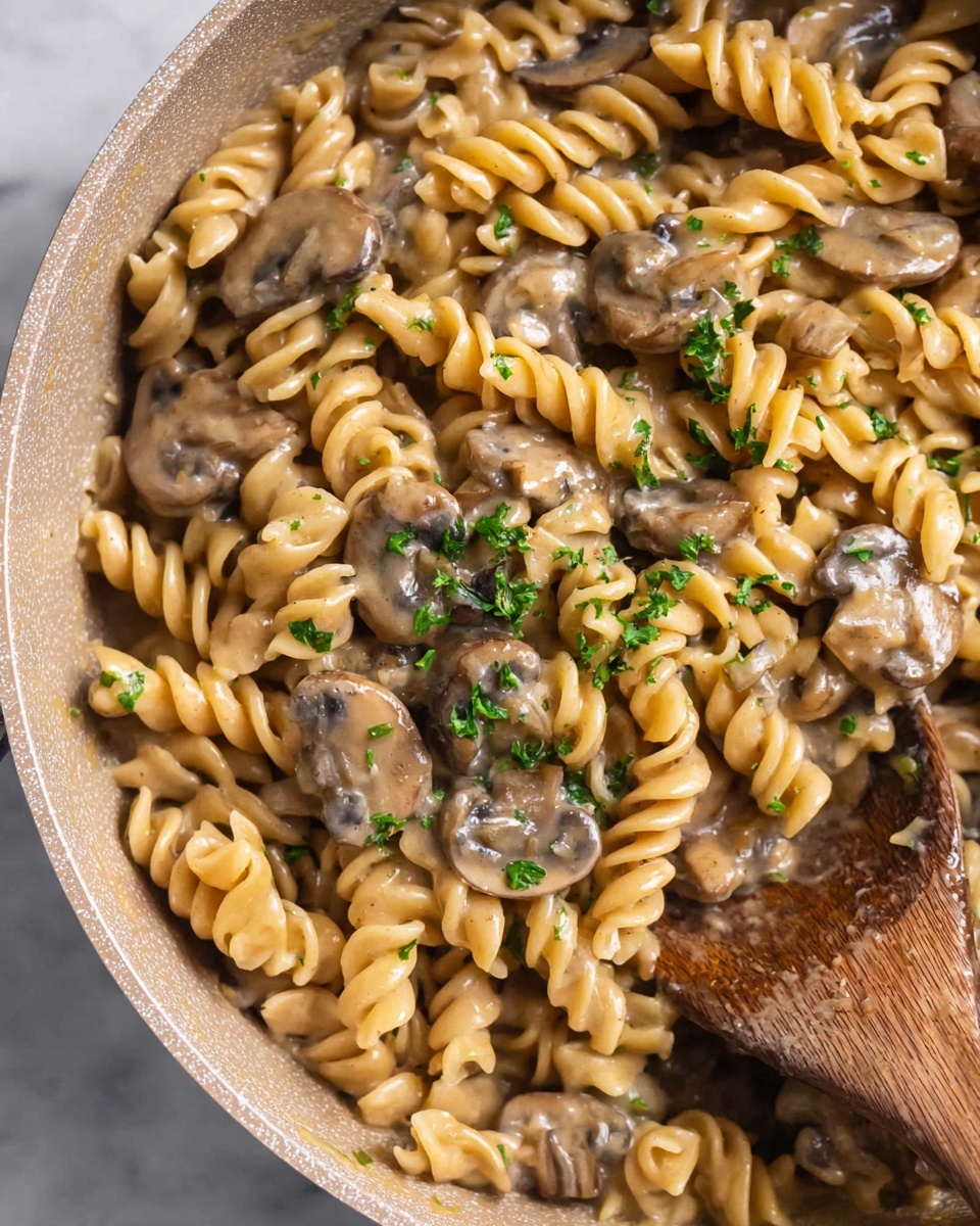 A close-up of a pan filled with creamy mushroom pasta made with spiral pasta, light brown sauce, and sliced cooked mushrooms. The pasta is well coated in the smooth sauce, which has a slightly shiny texture. Small green parsley pieces are sprinkled evenly on top, adding a touch of color. A wooden spoon is placed inside the pan, partially covered by the pasta. The pan itself has a soft beige color and a textured surface. The background is a white marbled texture photo taken with an iphone --ar 4:5 --v 7