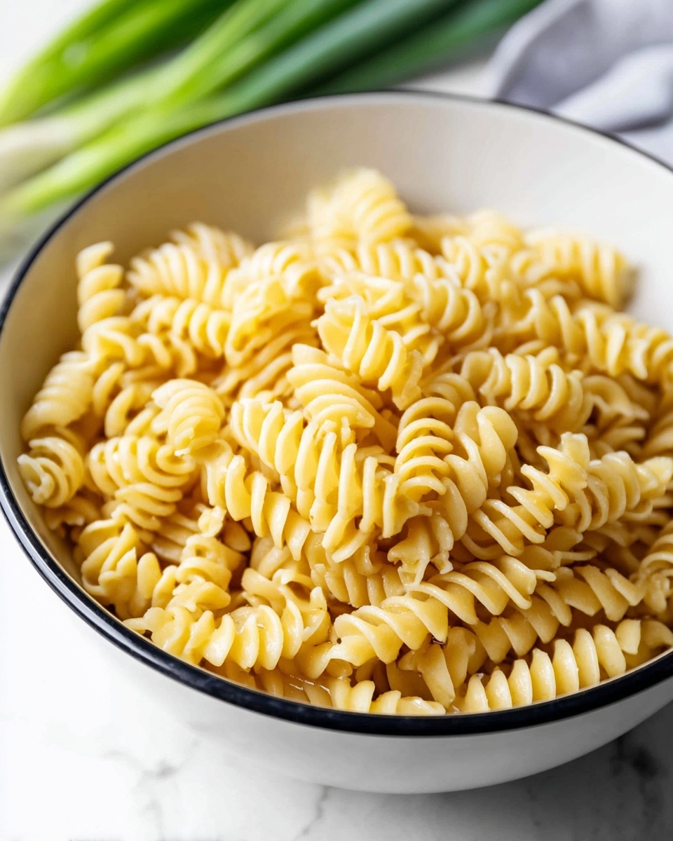 The image shows a white bowl with a black rim filled with cooked spiral pasta. The pasta has a light golden-yellow color and a slightly glossy texture, with tightly coiled pieces stacked unevenly inside the bowl. The bowl sits on a white marbled surface, and in the blurred background, there are green onions placed slightly out of focus. The lighting is bright and natural, emphasizing the pasta’s smooth and slightly firm appearance. photo taken with an iphone --ar 4:5 --v 7