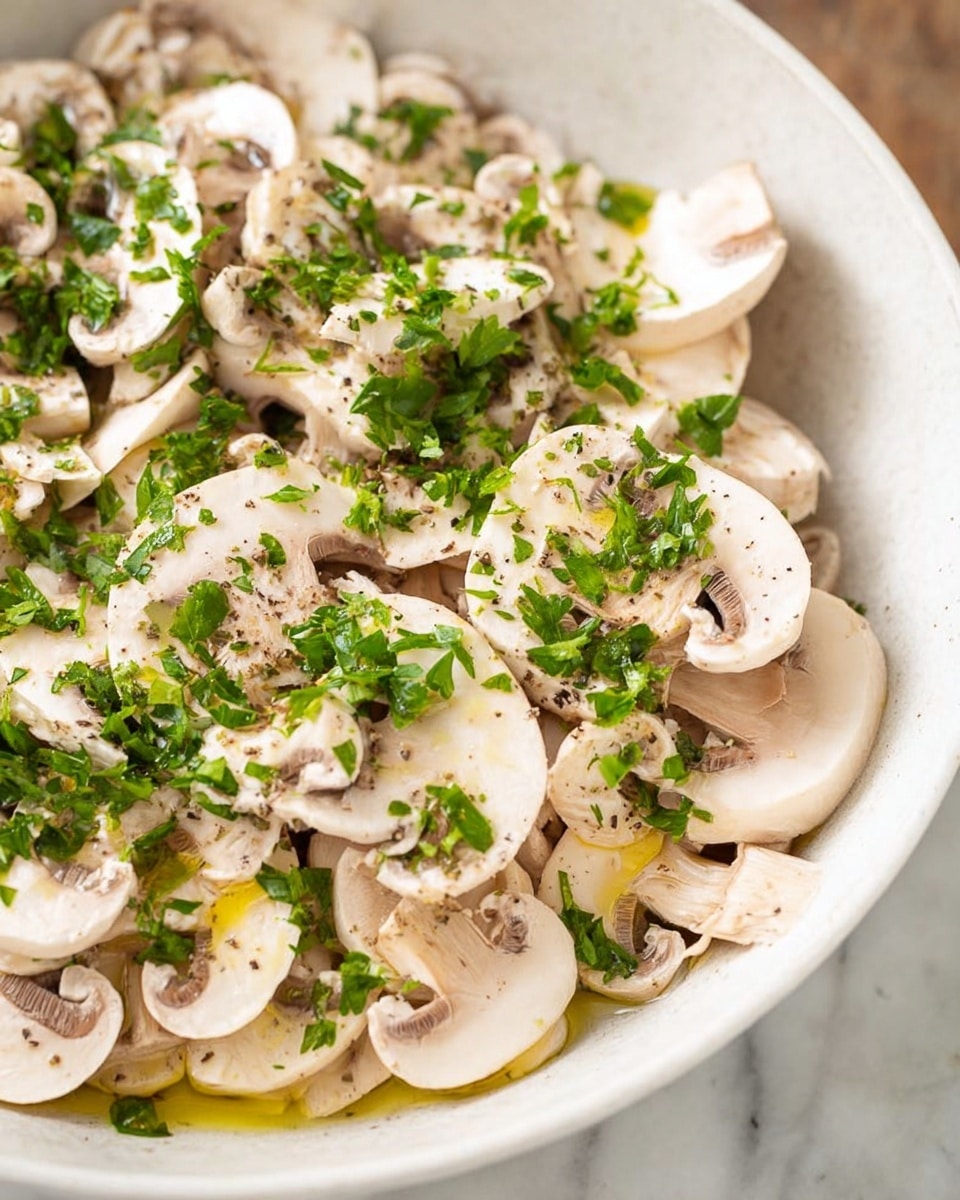 A close-up view of a white bowl filled with several layers of thinly sliced white mushrooms that have a smooth texture and light brown gills visible. On top of the mushroom slices, there is a sprinkling of finely chopped green herbs, likely parsley, which adds bright green color and a fresh look. A light drizzle of yellowish oil is visible over the mushrooms and herbs, giving a shiny and slightly wet appearance. The bowl is set on a white marbled surface. photo taken with an iphone --ar 4:5 --v 7