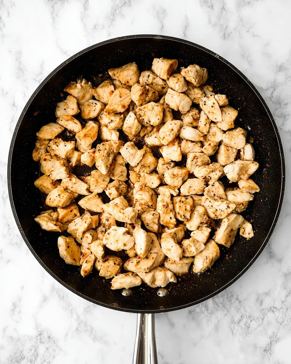 A round black pan filled with many pieces of light brown cooked chicken, each piece small and uneven in shape, showing light golden spots from cooking. The chicken pieces cover the whole pan, sitting close to each other. The pan rests on a white marbled surface, with a silver handle visible on the left side. photo taken with an iphone --ar 4:5 --v 7