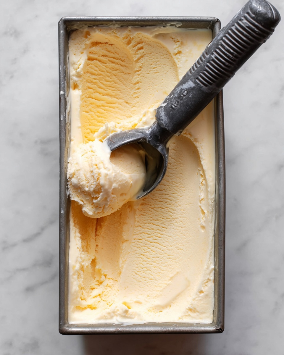 A rectangular metal container filled with creamy, pale yellow ice cream showing a soft, smooth texture with slight ridges on the surface. A dark gray ice cream scoop with a grooved handle rests on top, partially dipped into the ice cream, with some ice cream clinging to the scoop. The container sits on a white marbled surface. photo taken with an iphone --ar 4:5 --v 7