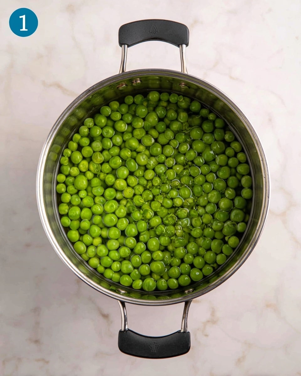 A top-down view of fresh green peas soaking in clear water inside a shiny silver metal pot with a black handle is shown. The peas cover the entire bottom layer of the pot, with the water level just above them. The pot rests on a white marbled surface, with a blue circle marked with the number