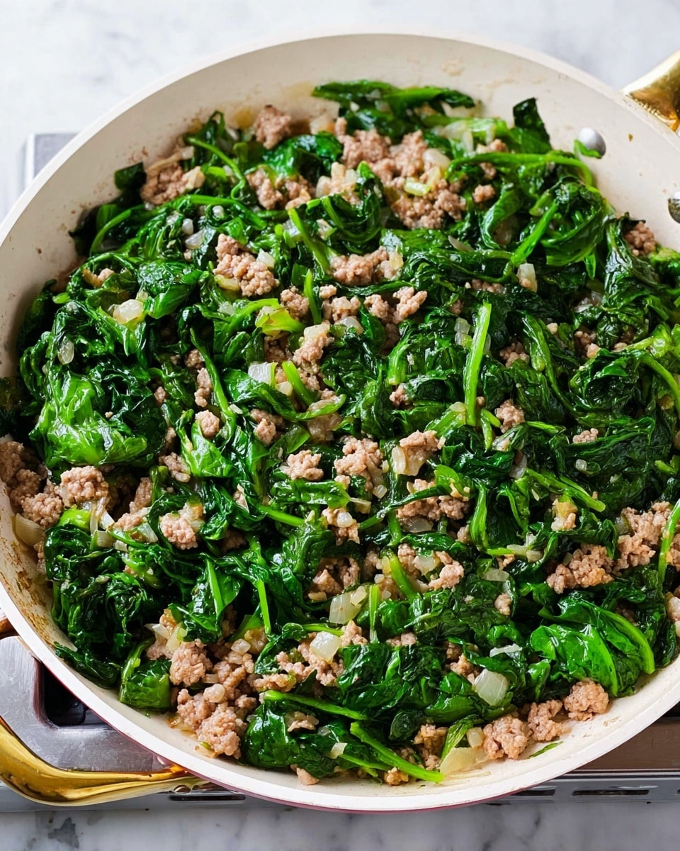 The image shows a white pan filled with cooked minced meat mixed with wilted bright green spinach leaves evenly spread throughout the pan. The meat is light brown and crumbly, sitting mainly under and between the spinach leaves. The white pan has gold handles, and the background is a white marbled texture with a stove edge visible under the pan. The mix looks fresh, warm, and lightly sauced with small bits of onion or garlic scattered within the dish photo taken with an iphone --ar 4:5 --v 7