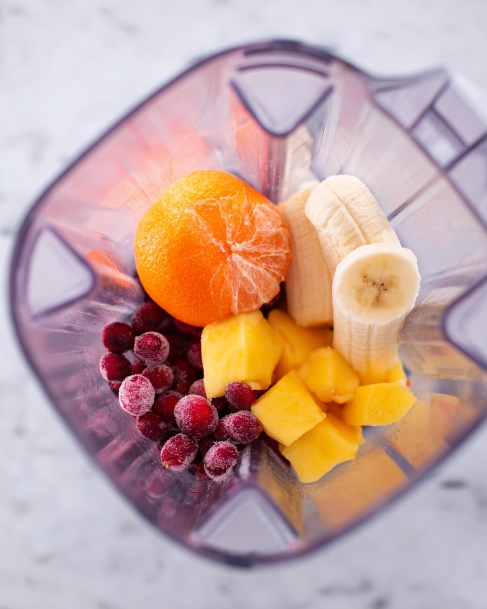 Inside a clear blender container, there are several layers of fruit. At the bottom, there is a mix of small red and purple round berries with a smooth, shiny texture. Above them is an unpeeled orange tangerine with a slightly rough surface and bright orange color. To the right of the tangerine, there are chunks of yellow mango with a soft, slightly fibrous texture. On the upper right side, a sliced banana with a pale yellow color and soft texture curves along the edge. The blender container is set against a white marbled textured surface. photo taken with an iphone --ar 4:5 --v 7