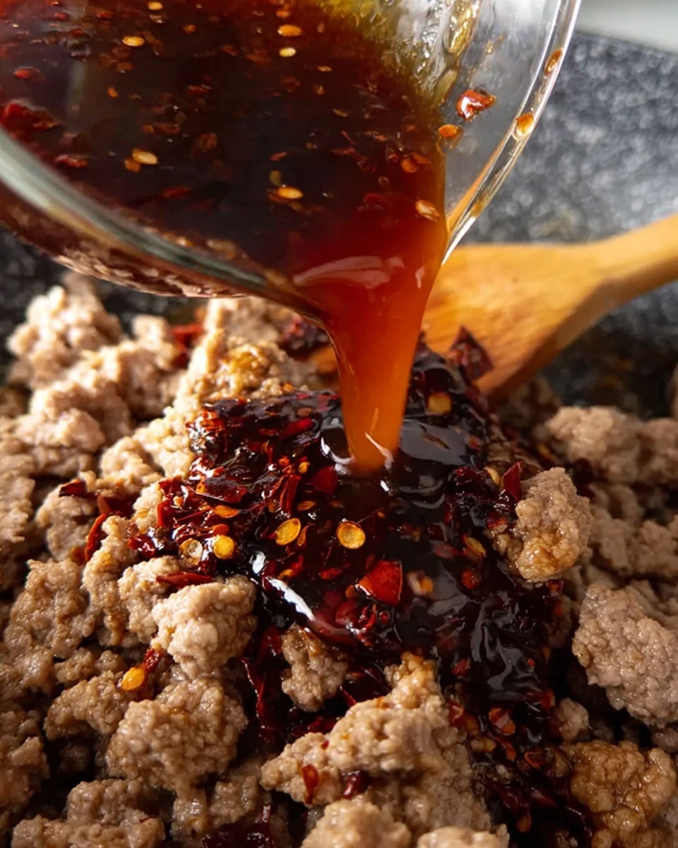 A close-up image shows dark brown sauce with red chili flakes being poured from a curved glass container onto a bed of light gray cooked ground meat in a pan. The sauce is thick and glossy, slowly dripping onto the meat that has a crumbly texture with small, uneven pieces. The focus is on the sauce stream and meat, with a wooden spoon partly visible on the right side. The background is out of focus, emphasizing the cooking action. photo taken with an iphone --ar 4:5 --v 7