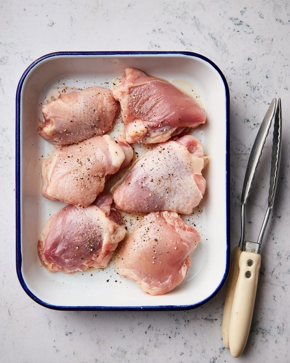 A white rectangular baking dish with a blue rim holds five raw pink chicken thighs. The chicken pieces are spread out evenly and lightly sprinkled with black pepper and salt. Next to the dish on the right are silver tongs with a beige handle resting on a white marbled surface. Photo taken with an iphone --ar 4:5 --v 7