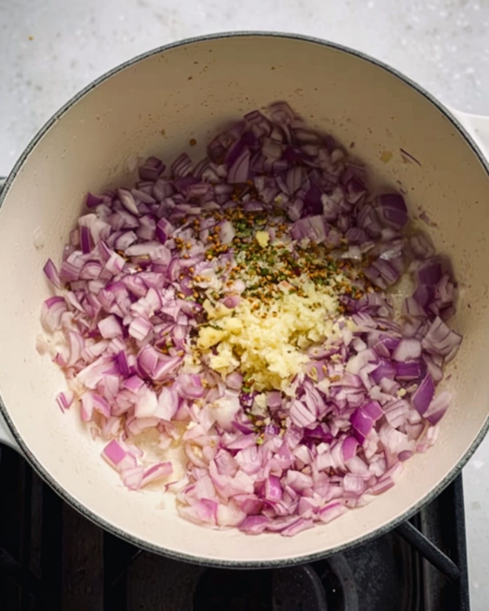 A close-up view shows a white pot on a white marbled surface filled with chopped red onions spread evenly over the bottom. In the center, there is a small pile of finely chopped fresh ginger, light yellow in color with a few small green bits mixed in. The onions have a slightly translucent texture, and you can see small seeds, likely cumin, scattered among them. The stovetop burner beneath the pot is visible but blurred. Photo taken with an iphone --ar 4:5 --v 7