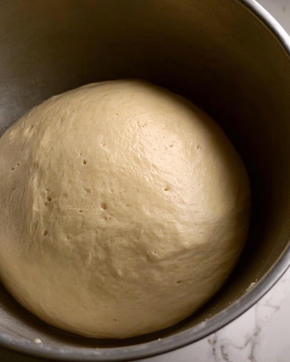 A close-up view of a single, smooth, light beige dough ball settled inside a large stainless steel bowl, with a slightly glossy surface showing small air bubbles, and the bowl placed on a white marbled texture. photo taken with an iphone --ar 4:5 --v 7