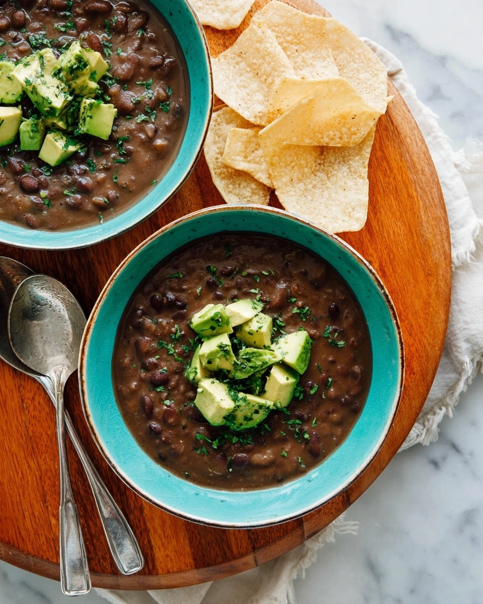 Two turquoise bowls filled with thick dark brown bean soup are shown from above. Each bowl has a layer of fresh, chunky avocado pieces in the center, sprinkled with chopped green herbs. The bowls rest on a round wooden board that has two silver spoons placed side by side. Pale tortilla chips are positioned near the top edges of the bowls. The background is a white marbled surface. photo taken with an iphone --ar 4:5 --v 7