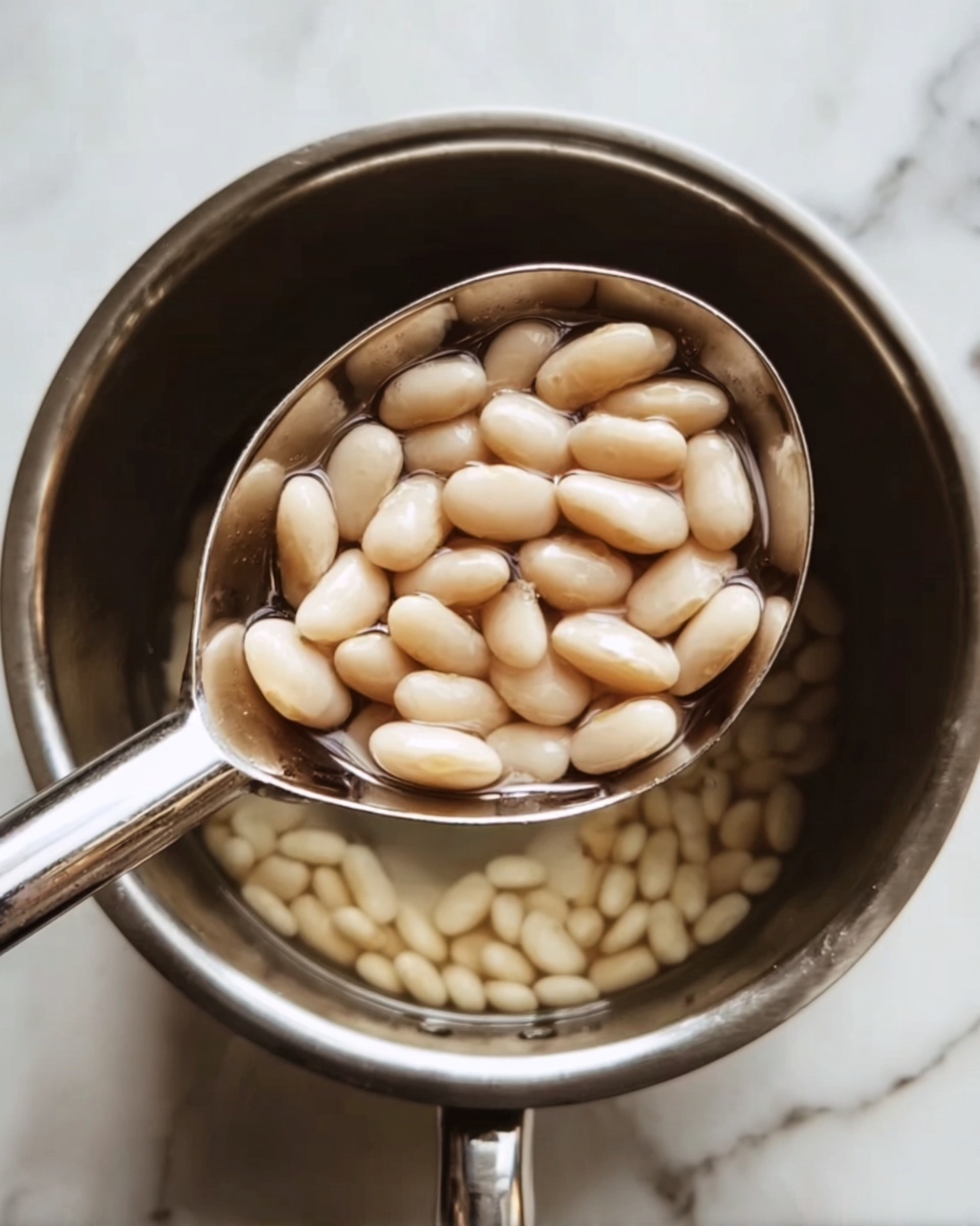 A close-up view of a silver ladle filled with small, smooth, light beige beans floating in clear water inside a stainless steel pot. The shiny ladle is held above the pot showing the beans clearly, with some beans visible at the bottom of the pot through the water. The background surface is a white marbled texture. Photo taken with an iphone --ar 4:5 --v 7