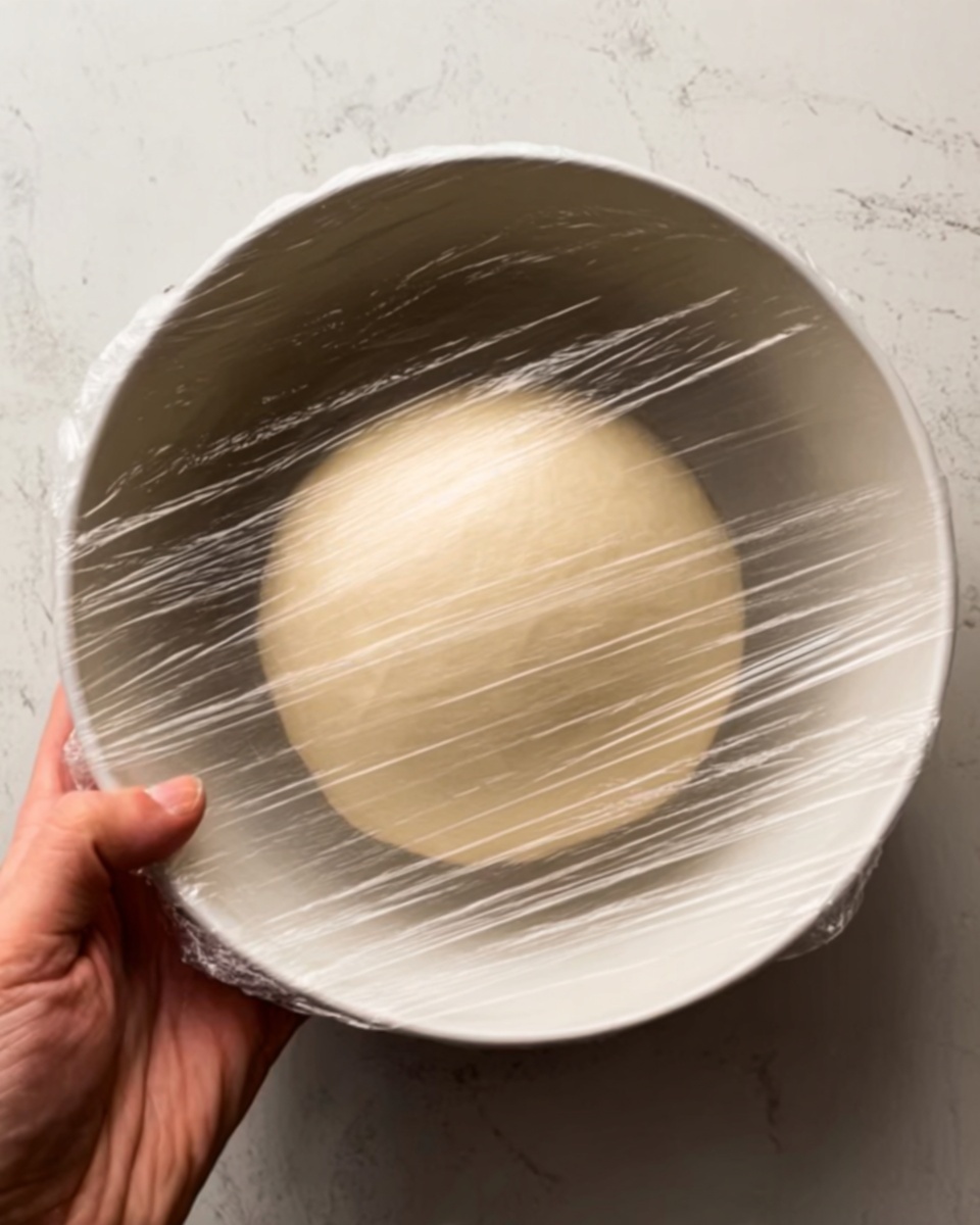 The image shows a single round dough ball with a smooth, pale beige surface, resting inside a large white bowl. The dough is covered tightly with clear plastic wrap that creates slight reflections and wrinkles on its surface. The woman’s hand supports the bowl from the bottom left corner, with fingers gently holding it. The background is a white marbled texture, visible around the edges of the bowl. photo taken with an iphone --ar 4:5 --v 7