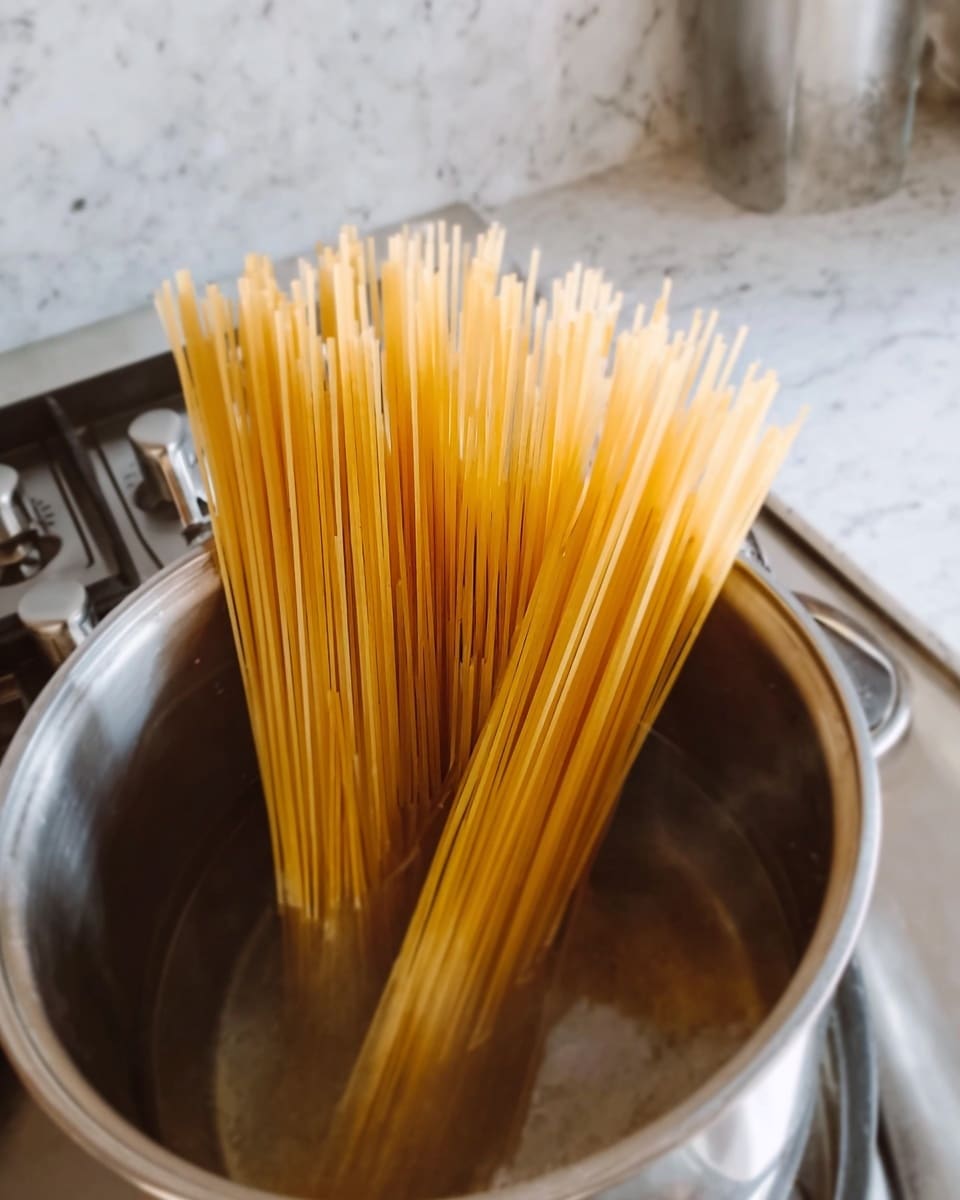 A close-up of a silver pot filled with clear water, sitting on a stove with metal control buttons in the background. Inside the pot are many long, uncooked spaghetti noodles standing upright, partially submerged in the water with the top ends still dry and sticking out above the pot's rim. The stove has a white marbled surface beneath the pot. Photo taken with an iphone --ar 4:5 --v 7