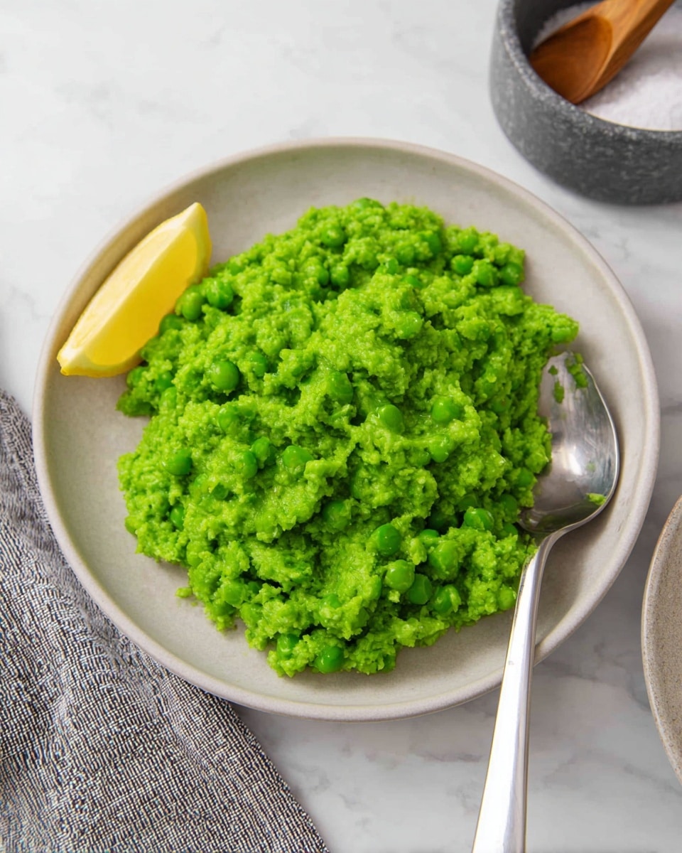 A mound of bright green mashed peas sits in the center of a white plate, showing a soft and slightly chunky texture. The green mash fills most of the plate’s surface, with a shiny silver spoon placed on the right side, partially under the mash. A fresh lemon wedge with a glossy yellow peel rests on the left side of the plate, adding a pop of color. The plate is set on a white marbled surface, with a gray and white striped cloth visible in the bottom corner and a round gray salt container with a wooden spoon in the background. Photo taken with an iphone --ar 4:5 --v 7