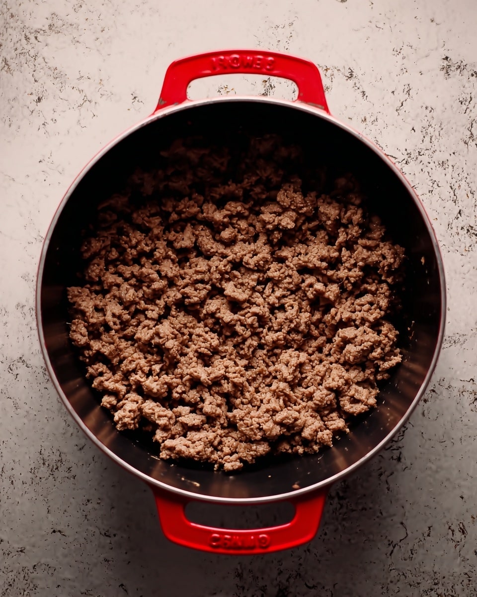 A close-up top view of cooked ground beef inside a large black pot with bright red handles on each side. The beef is broken into small, crumbly pieces with a slightly brown color and a dry texture. The pot is placed on a surface with a white marbled texture. photo taken with an iphone --ar 4:5 --v 7