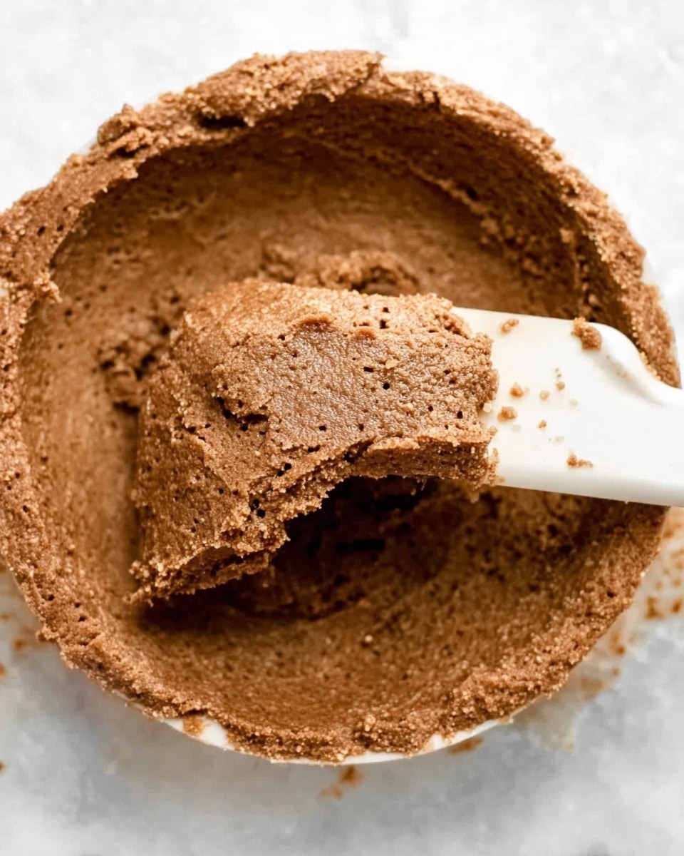 A close-up image of thick, rough-textured brown dough being spread in a low round container with a white spatula, showing a slightly uneven surface with visible tiny holes and specks. The dough is dense and appears moist, with a portion scooped away revealing the smooth inner side. The container is placed on a white marbled texture surface with some stray bits of dough around it. Photo taken with an iphone --ar 4:5 --v 7