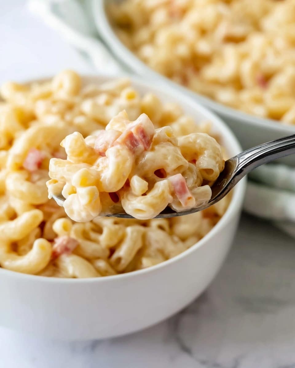 A close-up of a spoon full of creamy macaroni pasta with small pieces of pinkish ham mixed in, showing a smooth texture with the sauce coating the pasta well. In the background, a white bowl filled with the same macaroni mixture is partially visible, sitting on a white marbled surface. The macaroni is light yellow with a creamy finish and the bits of ham add a touch of soft pink throughout the dish. Photo taken with an iphone --ar 4:5 --v 7