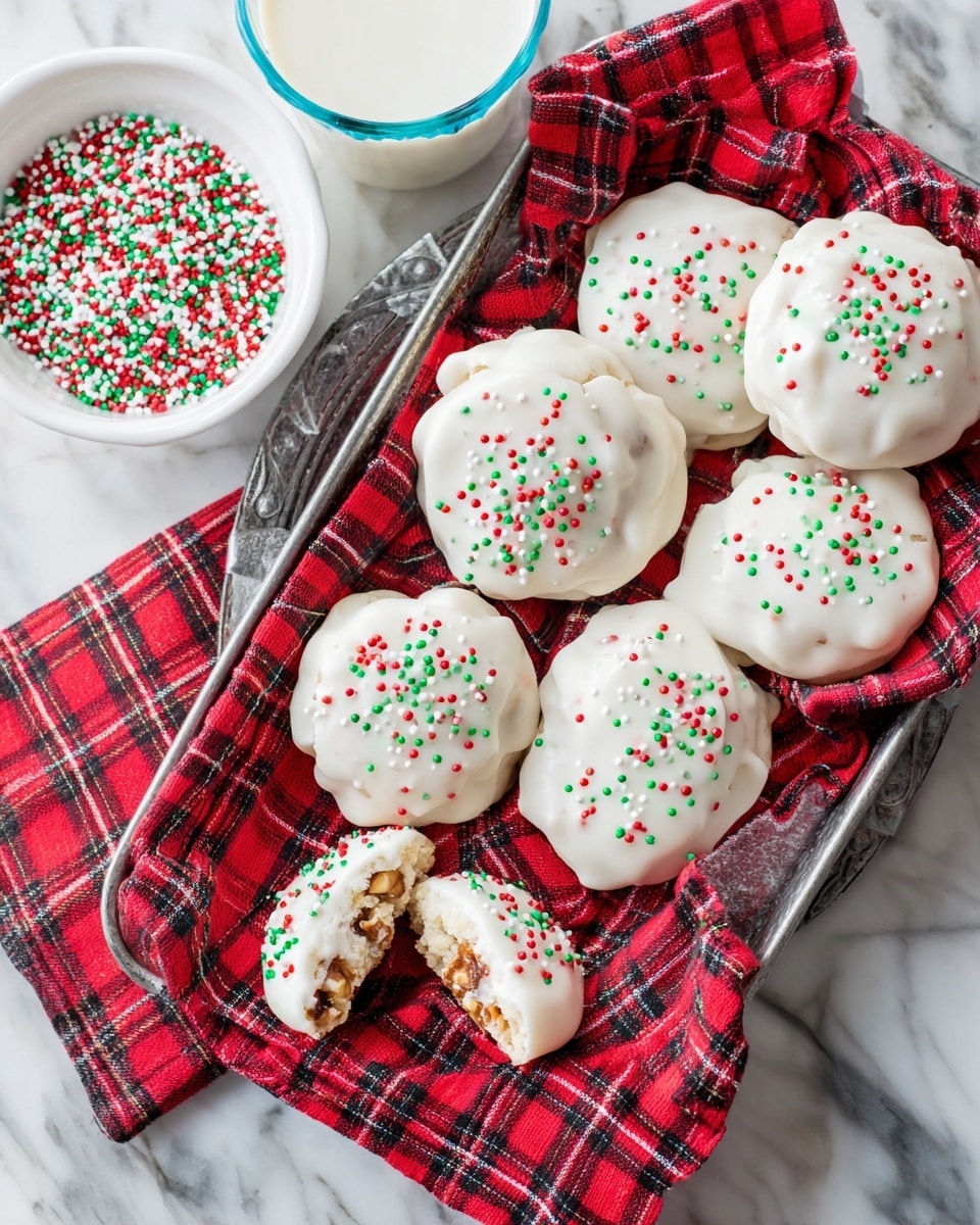 A metal tray lined with a red and black checkered cloth holds around ten round white cookies covered in white frosting, each topped with small red, green, and white round sprinkles. One cookie is cut in half showing a layer of nuts inside under the frosting. Next to the tray, there is a small white bowl filled with the same sprinkles and a glass of milk with a blue rim. The background is a white marbled surface. Photo taken with an iphone --ar 4:5 --v 7