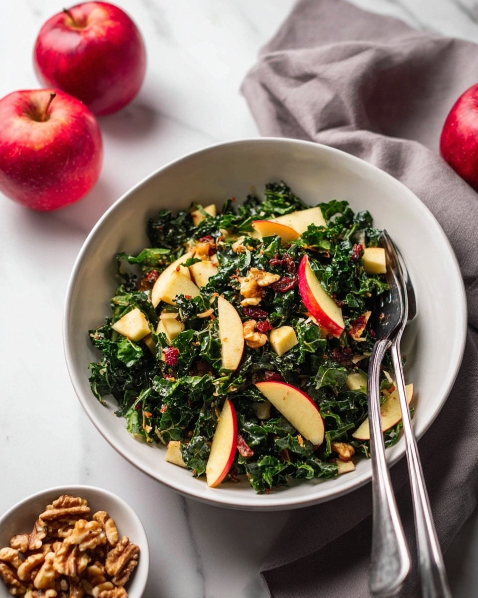 A white bowl filled with a kale salad that has three main layers: shiny dark green kale leaves forming the base, light yellow-green apple slices with red skin mixed throughout, and small light brown walnut pieces scattered on top. Two silver forks rest inside the bowl on the right side. The bowl sits on a white marbled surface next to two red apples in the front left and a small white bowl of walnuts in the background. A soft gray cloth is partially visible behind the salad bowl. photo taken with an iphone --ar 4:5 --v 7