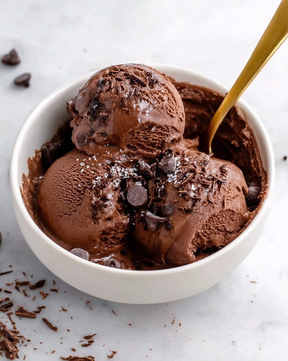 A white bowl filled with three large scoops of rich, creamy dark chocolate ice cream, slightly melting and with a shiny surface. The ice cream has small chocolate bits inside and is topped with fine shavings of chocolate scattered across the top. The bowl sits on a white marbled surface with a few chocolate chips and crumbs around it. A golden spoon is placed inside the bowl, partially covered by the ice cream. Photo taken with an iphone --ar 4:5 --v 7