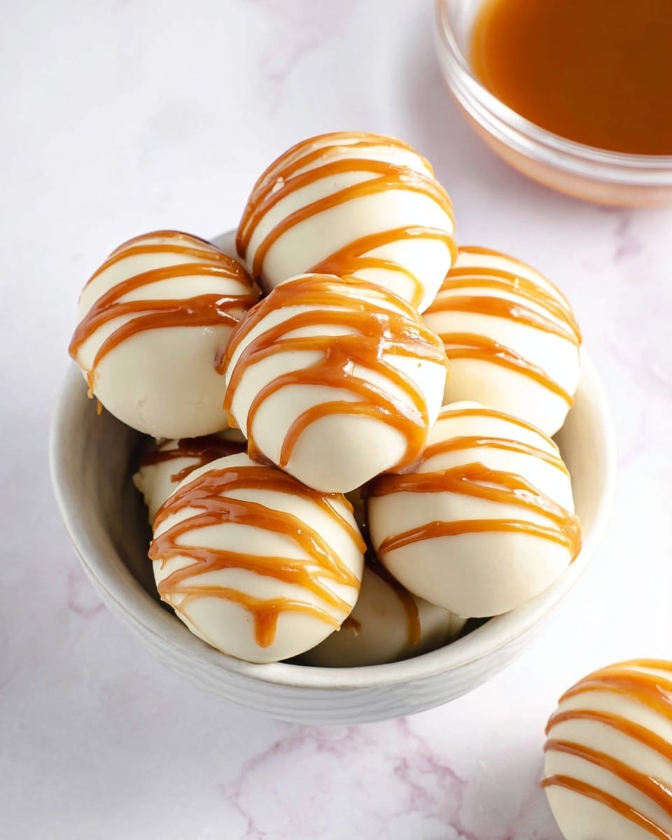 The image shows a white bowl holding several round white balls that look smooth and creamy. Each ball has three diagonal caramel-colored stripes drizzled on top, giving them a shiny and slightly thick look. The balls are neatly stacked, filling the bowl almost to the top. In the top left corner, part of a clear bowl with light brown caramel sauce is visible, and in the top right corner, another ball with the same caramel stripes is partially shown. The background surface is a white marbled texture. photo taken with an iphone --ar 4:5 --v 7