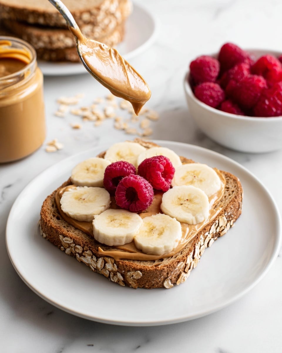 The image shows a single slice of brown oat bread on a white plate with visible oats on the crust. The first layer on the bread is a smooth peanut butter spread evenly. On top of the peanut butter, there are five slices of banana laid out in a circle near the center of the bread. Centered on the banana slices are three whole fresh raspberries, bright red and plump. Above the raspberries, a spoon with creamy peanut butter is held, with some peanut butter about to drop onto the berries. In the background, there is a white bowl with more raspberries, a jar of peanut butter, and a stack of similar oat bread slices, all on a white marbled surface photo taken with an iphone --ar 4:5 --v 7
