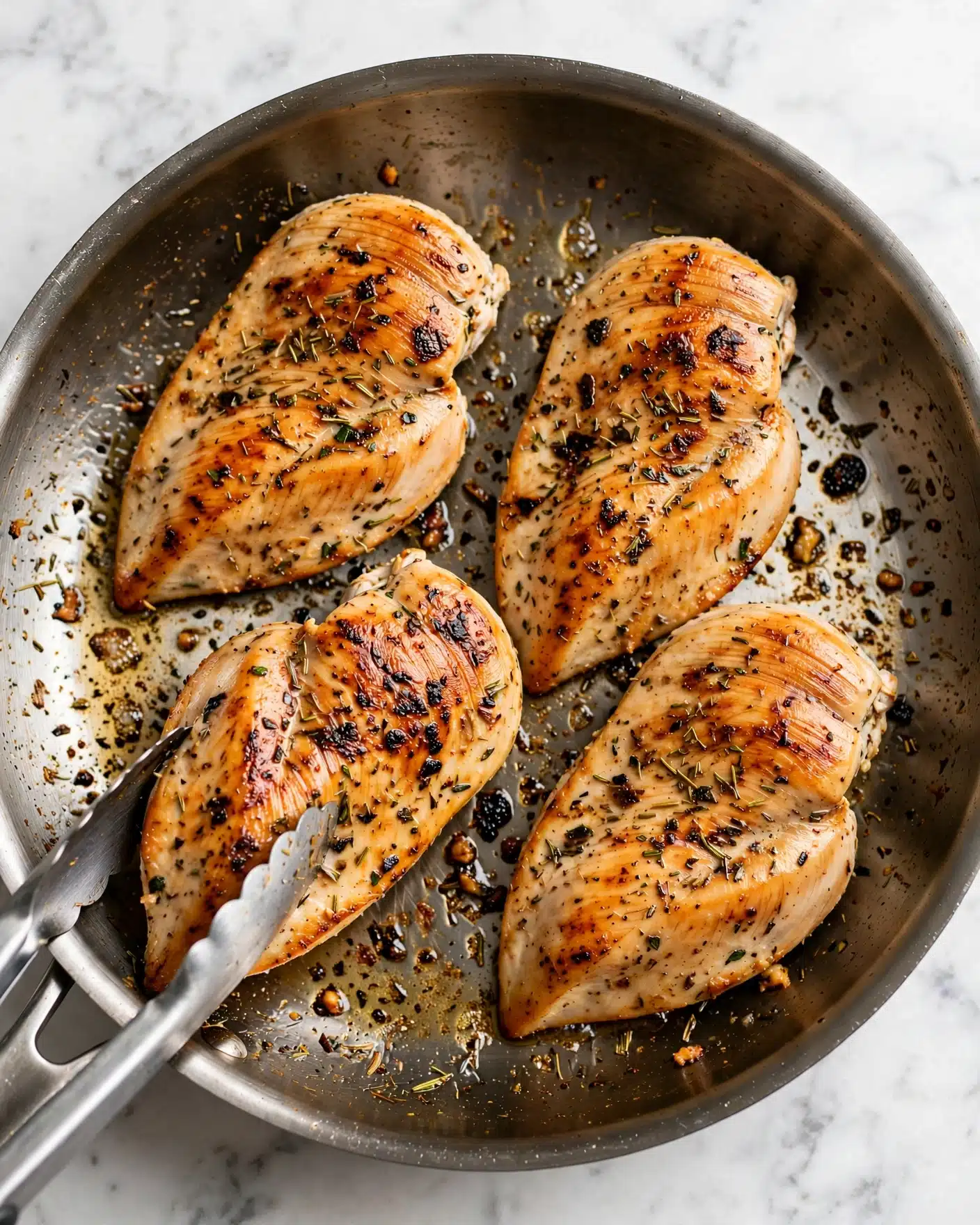 Creamy Garlic Chicken and Broccoli Recipe 4 The image shows four pieces of cooked chicken on a white skillet, each chicken piece golden brown with some darker brown spots and sprinkled with dried herbs. The chicken pieces are placed close to each other, with one piece being held by tongs near the bottom left corner. The skillet has browned oil and small food bits around the chicken, enhancing the look of a hot pan. The skillet is on a white marbled surface. photo taken with an iphone --ar 4:5 --v 7