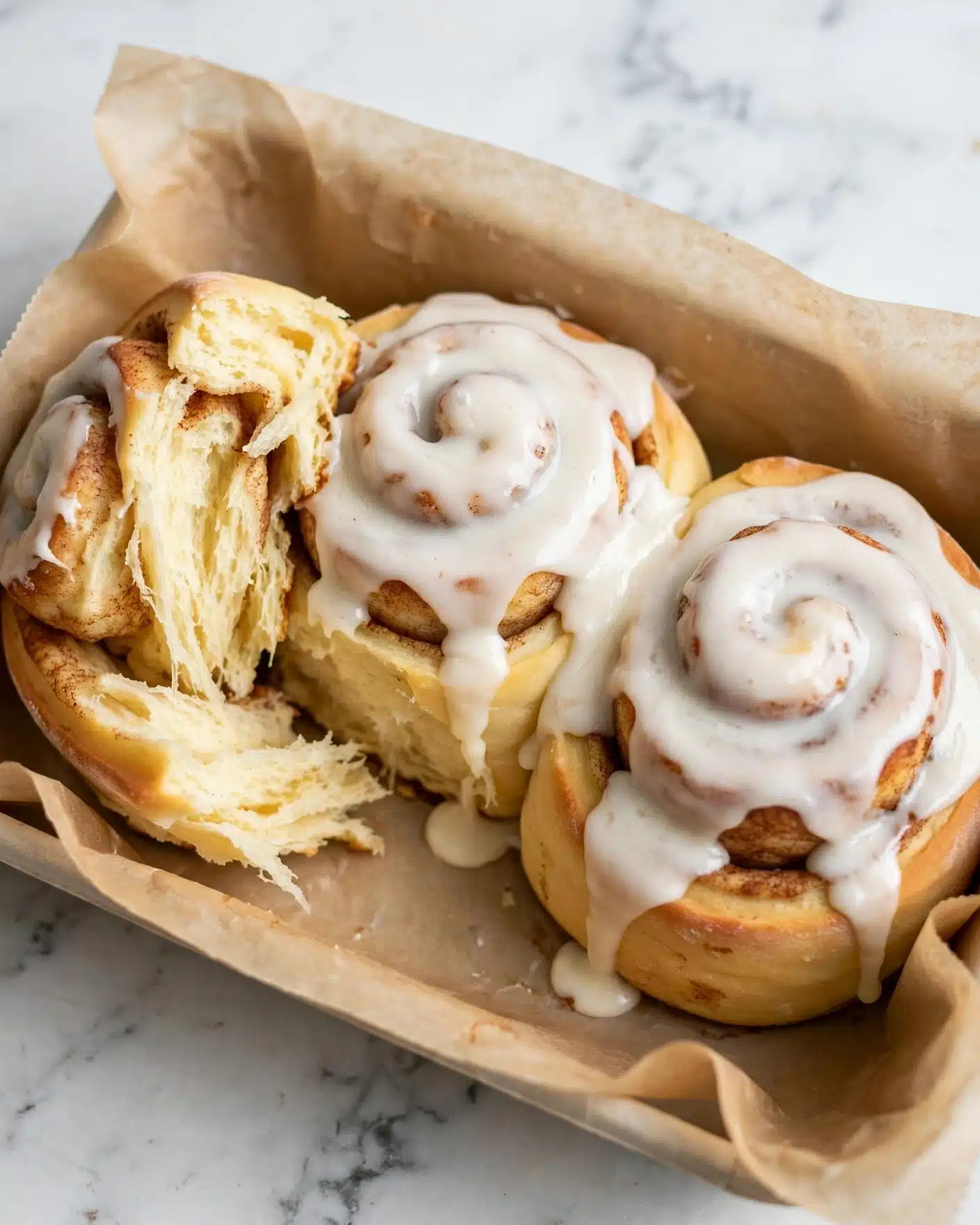 The image shows three soft cinnamon rolls placed close together in a container lined with light brown parchment paper. Each roll has a thick layer of white icing dripping over the top, with the dough underneath that is golden yellow and fluffy. One cinnamon roll is partially torn, showing the soft and airy inside texture with a slight yellow tint. The cinnamon rolls have swirled layers creating a spiral effect, with hints of cinnamon peeking through. The background is a white marbled texture. photo taken with an iphone --ar 4:5 --v 7