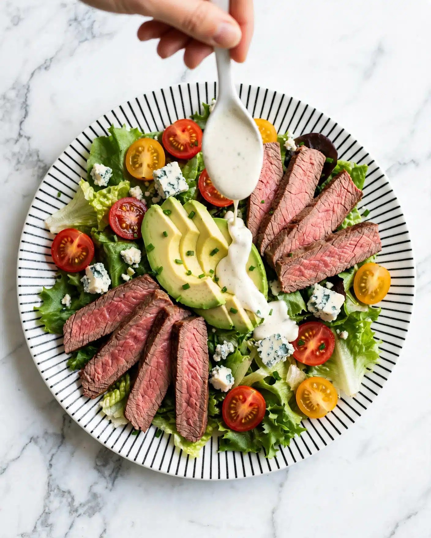 This image shows a salad on a white plate with black stripe patterns on the edge, placed on a white marbled surface. The salad has two main layers: at the bottom, there is a bed of green lettuce leaves with sliced yellow and red grape tomatoes scattered around. On top, there are five pieces of medium rare steak sliced thinly, arranged in a fan shape on opposite sides of the plate. Between the steak slices, there are thick slices of avocado with a pale green color. Some crumbled blue cheese and green chive pieces are scattered on top. A woman's hand holds a white spoon pouring a creamy white dressing over the salad. photo taken with an iphone --ar 4:5 --v 7