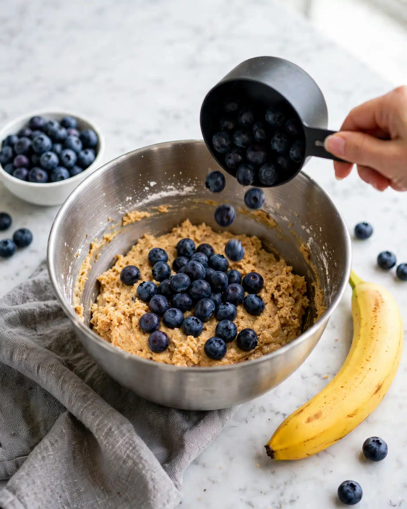 A silver mixing bowl sits on a white marbled surface, filled halfway with light brown batter that has a rough texture. On top of the batter is a loose pile of dark blue blueberries with a smooth, shiny appearance. A woman's hand holds a black measuring cup pouring more blueberries into the bowl, some mid-air. Nearby on the marble surface, there is a small white bowl full of blueberries to the left and a bright yellow banana to the right. A gray cloth lies partially visible under the mixing bowl. The scene is bright and clear, showcasing the fresh ingredients and mixing process. photo taken with an iphone --ar 4:5 --v 7
