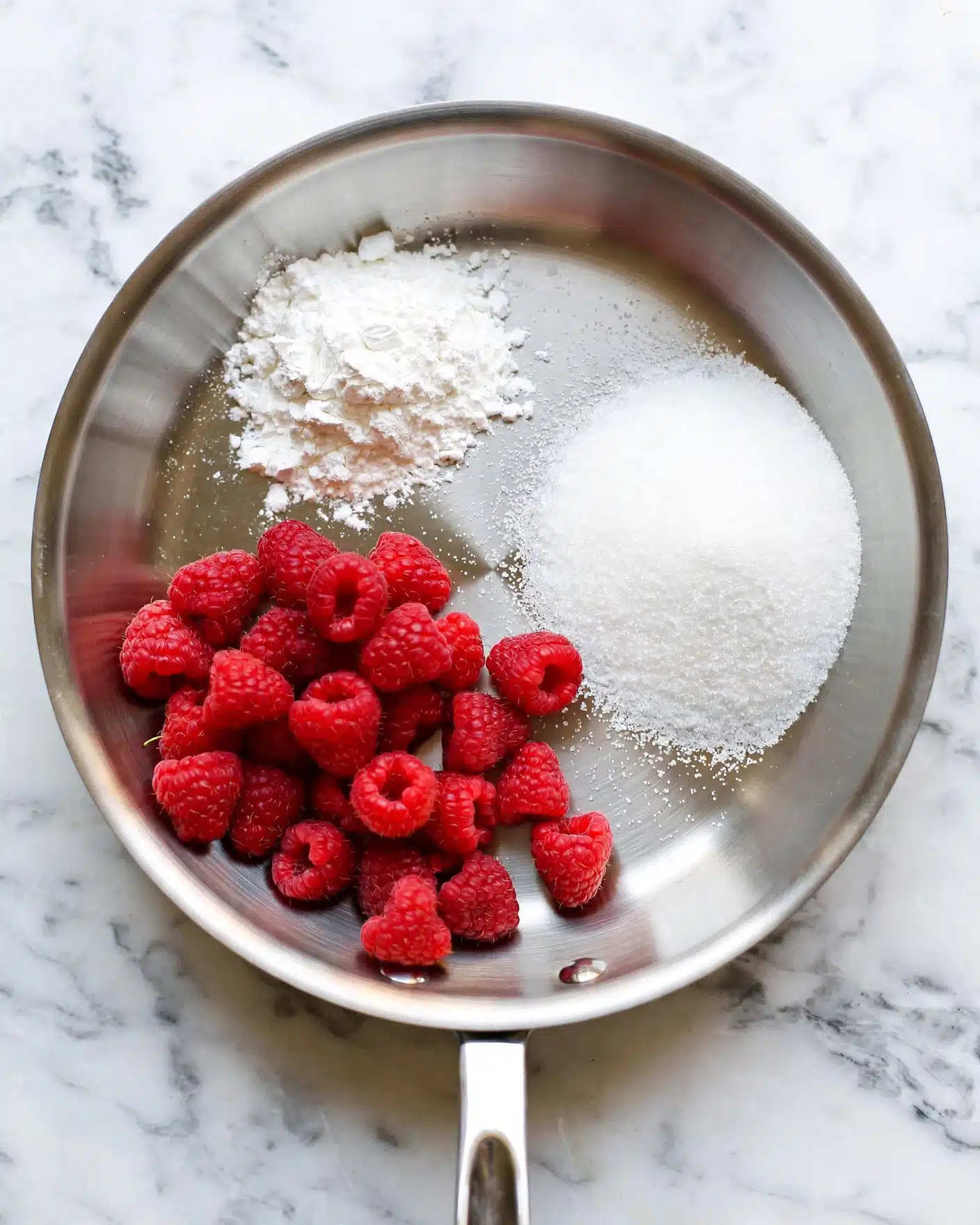 A round stainless steel pan sits on a white marbled surface, filled with three groups of ingredients each forming a separate section: bright red raspberries clustered at the bottom left, a small pile of white powdery cornstarch at the top left, and a mound of white granulated sugar on the right side. The metal pan has a smooth shiny texture and a short handle extending downwards. The colors contrast clearly, with the red raspberries standing out against the white powders and reflective metal photo taken with an iphone --ar 4:5 --v 7