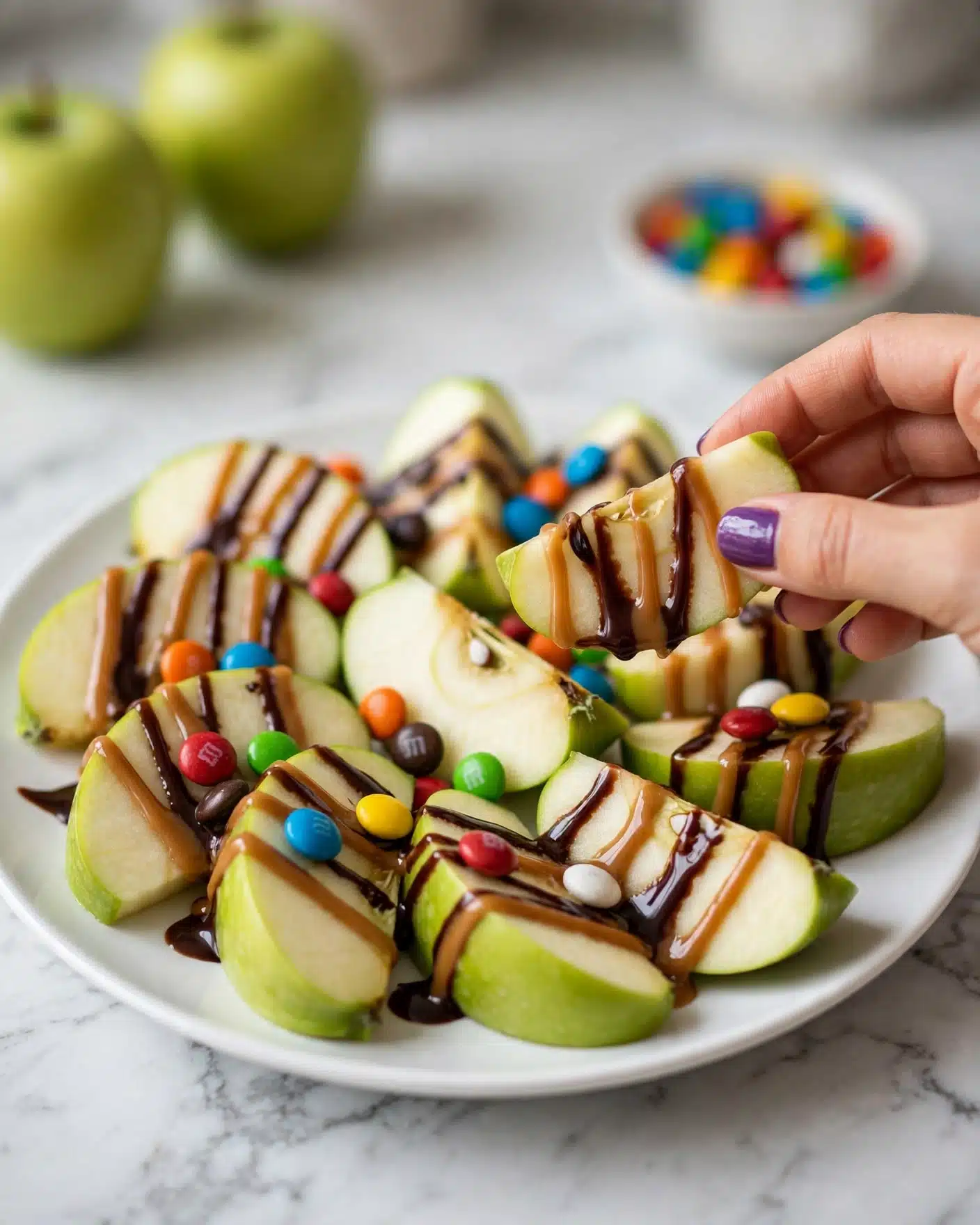 A white plate is filled with green apple wedges arranged in a slightly overlapping circle. The apple slices have a fresh, light yellow inside and bright green skin on the edges. Each wedge is drizzled with a layered mix of light brown caramel sauce and dark chocolate syrup, creating shiny lines over the fruit. Colorful candy pieces in blue, red, orange, yellow, green, brown, and white are scattered evenly on top of the apples and sauces. A woman's hand with purple nail polish picks up one wedge from the plate. The plate sits on a white marbled surface, and some green apples and a white bowl with more candies are blurred in the background. Photo taken with an iphone --ar 4:5 --v 7