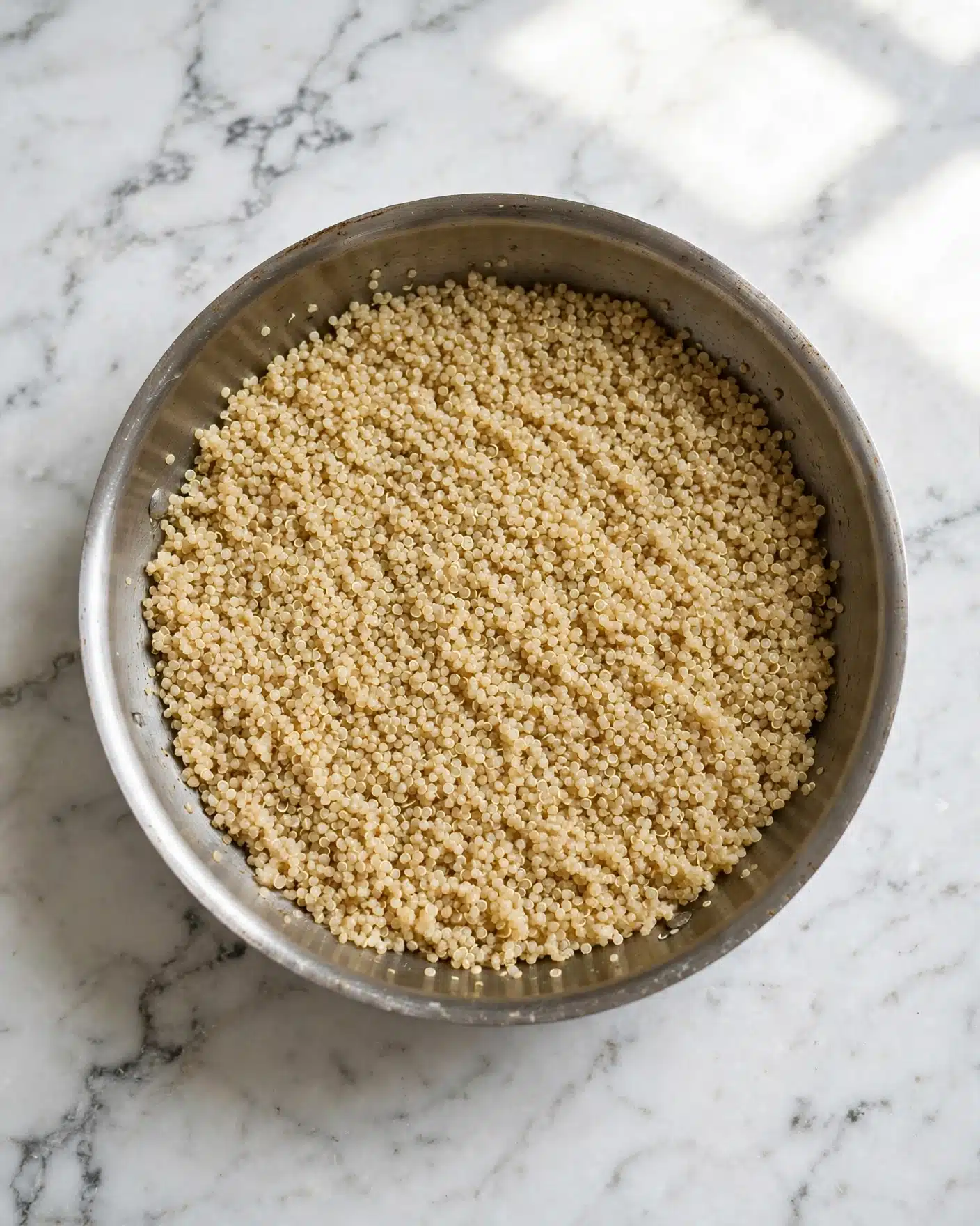 A metal pan with a smooth brown handle is filled with cooked quinoa. The quinoa grains look soft and fluffy, forming a textured light golden-yellow layer that almost fills the pan. The pan rests on a white marbled surface with a clean, subtle pattern. photo taken with an iphone --ar 4:5 --v 7