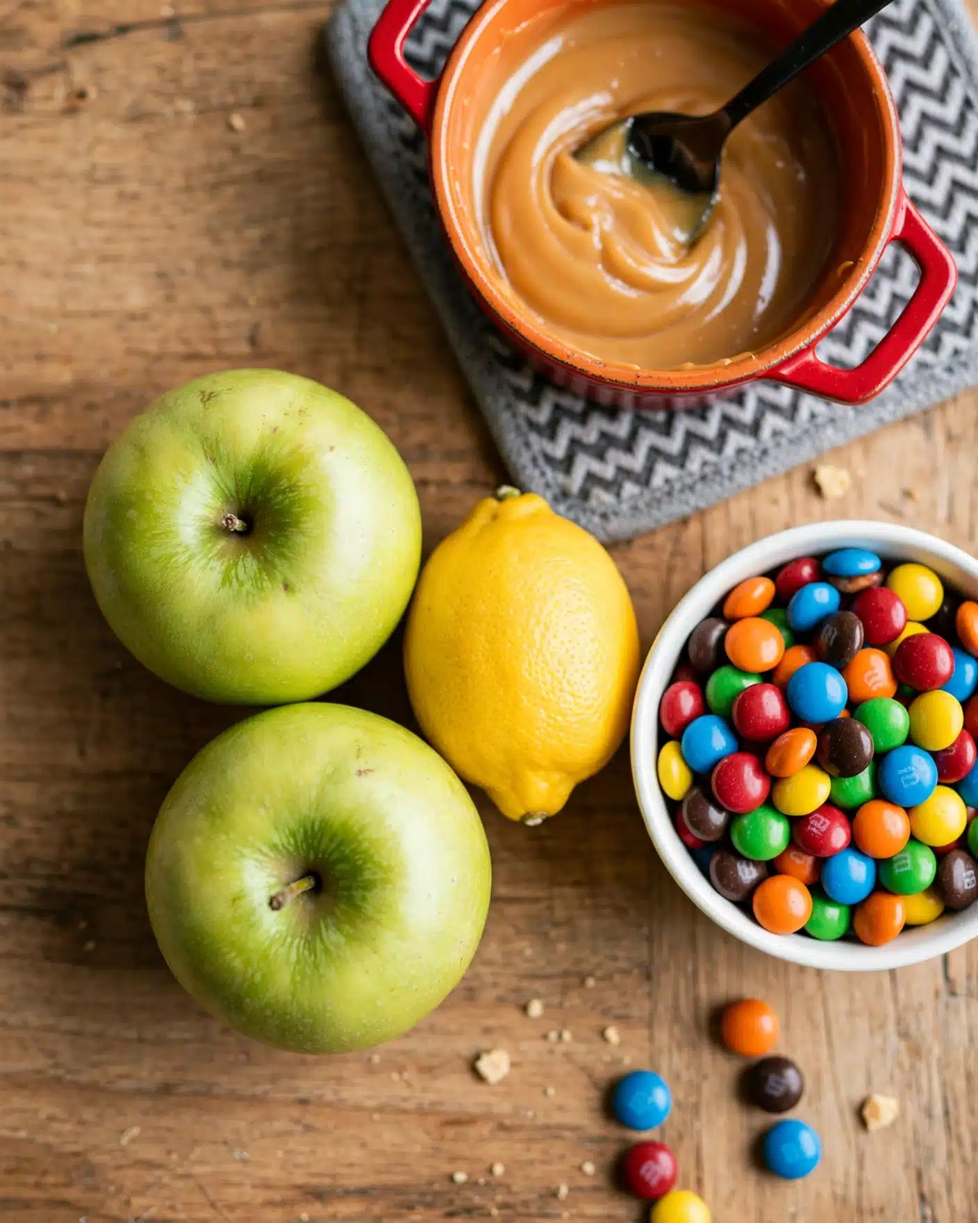 The image shows a top view of ingredients on a wooden surface, featuring two green apples and a bright yellow lemon placed close together near the center. To the right, there is a white bowl filled with small, colorful candy-coated chocolates in red, blue, green, orange, yellow, and brown colors. Above these, a red pot with an orange interior holds a smooth, creamy caramel-colored sauce with a black spoon resting inside, stirring the sauce gently. The pot sits on a gray trivet with a zigzag pattern. photo taken with an iphone --ar 4:5 --v 7