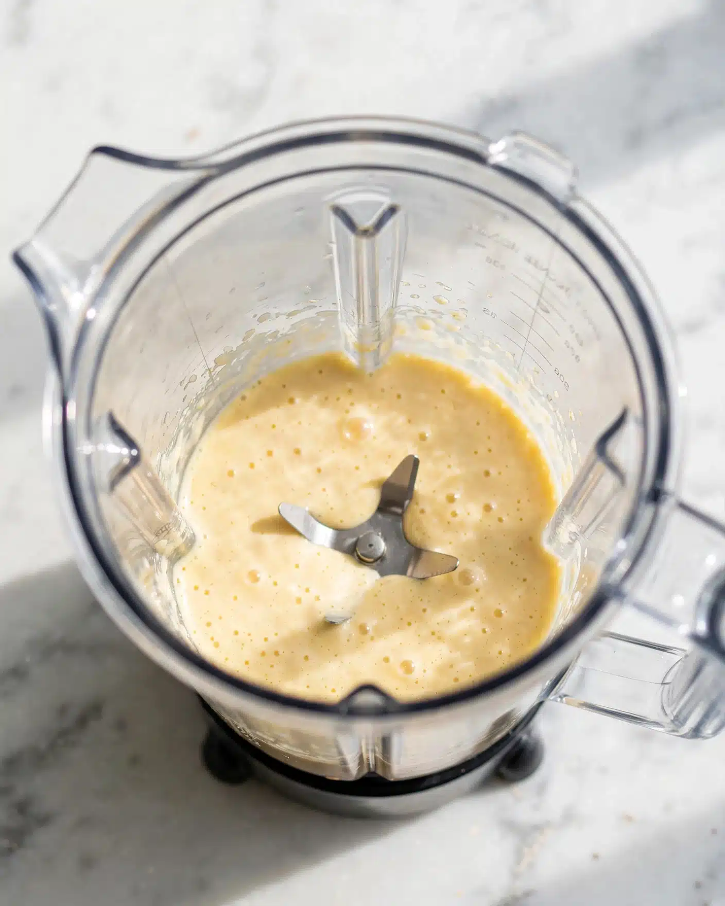 A clear blender seen from above shows a single layer of thick, light yellow creamy mixture with a smooth texture and a few small air bubbles sitting inside the blender container, which rests on a white marbled surface photo taken with an iphone --ar 4:5 --v 7