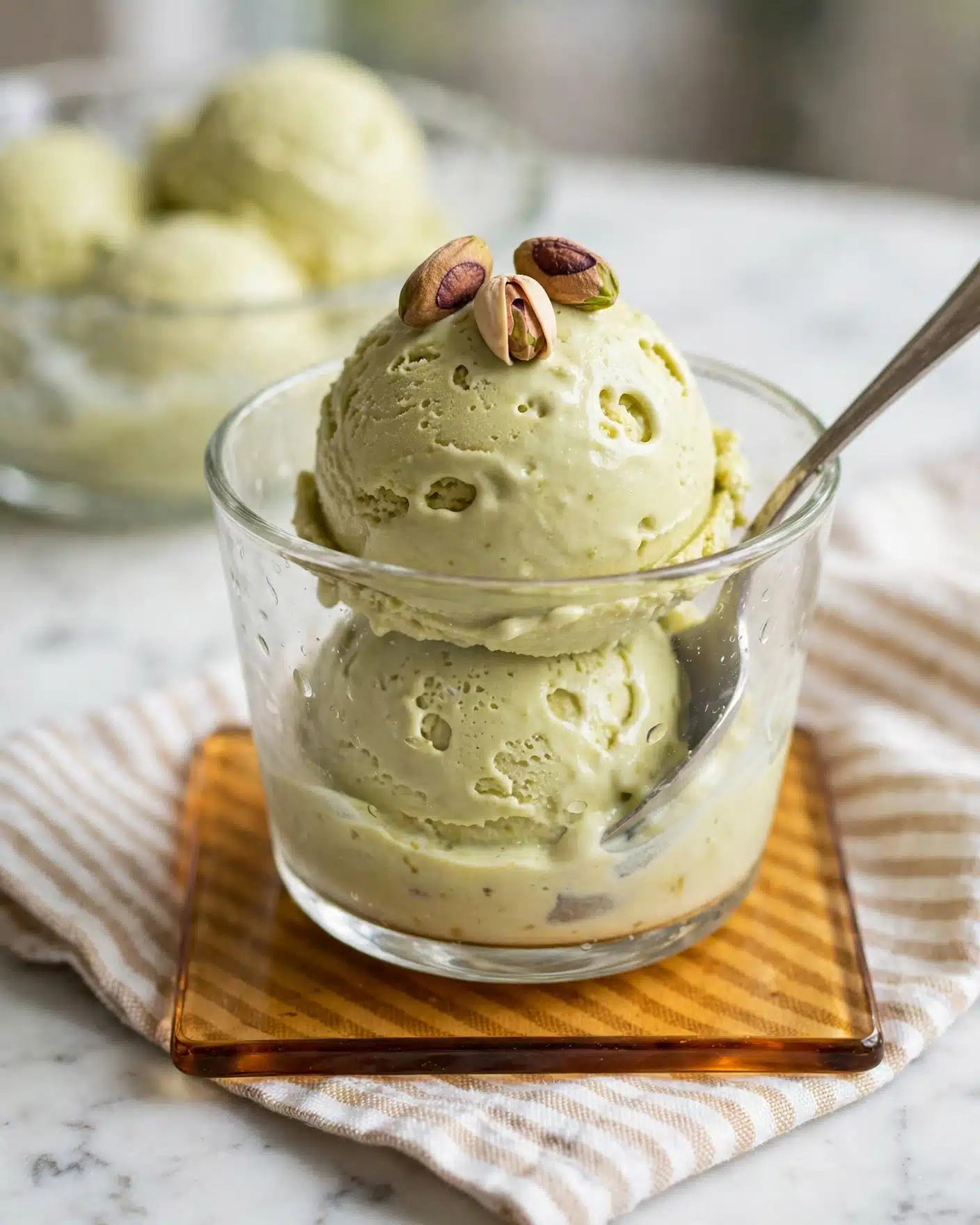 A clear glass cup holds two scoops of creamy pistachio-colored ice cream, one scoop stacked neatly on top of the other. The top scoop is decorated with three whole pistachio nuts showing green and brown shades. A silver spoon rests inside the cup, surrounded by the smooth texture of the ice cream. The glass cup sits on a square glass coaster with amber tones, placed on a white marbled surface with a striped cloth underneath. In the blurred background, there is a glass dish filled with more of the same ice cream. Photo taken with an iphone --ar 4:5 --v 7