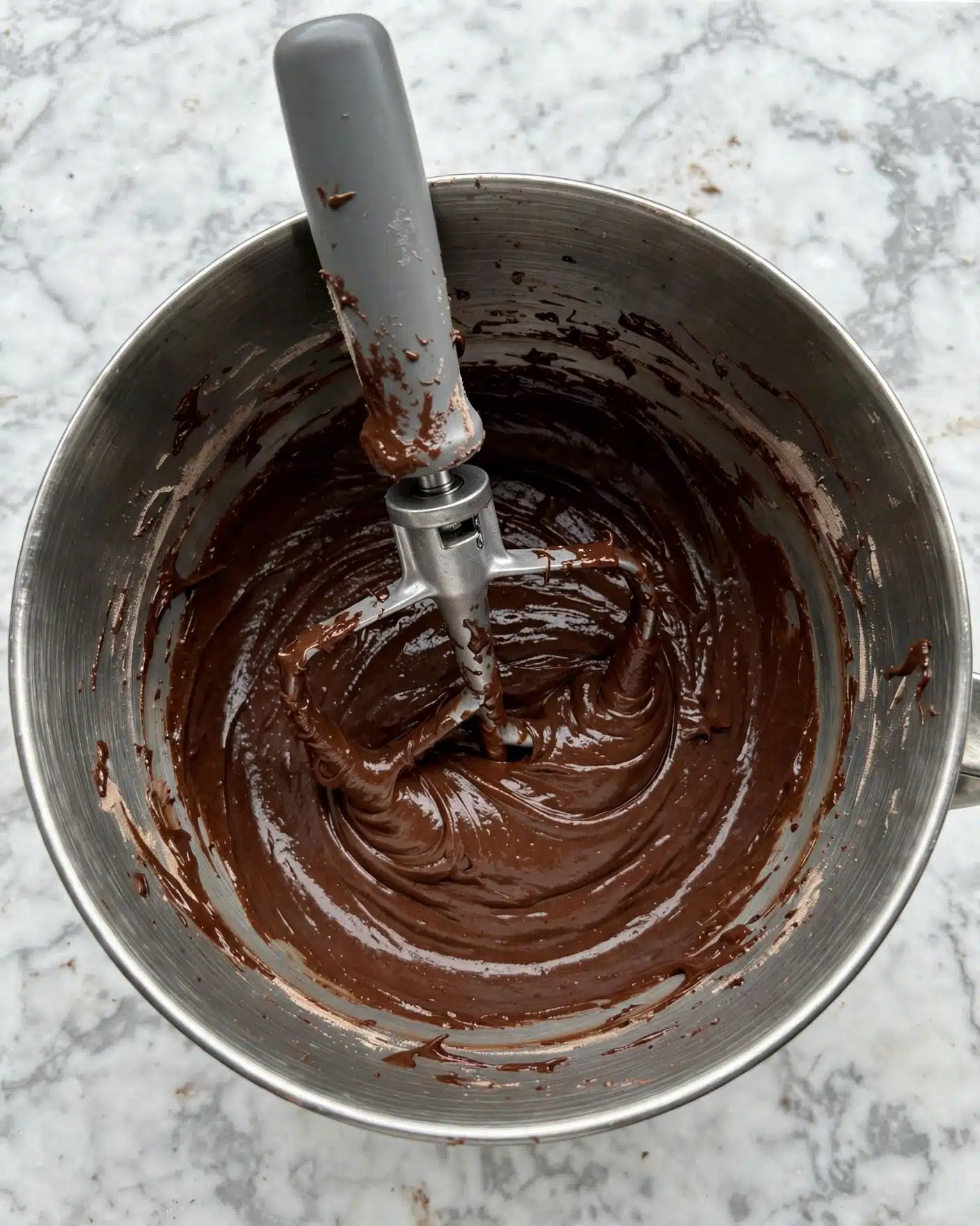 Espresso Martini Cookies Recipe 4 A top-down view of a shiny metal bowl filled with thick, dark brown chocolate batter that has a smooth and dense texture. Inside the bowl, a metal mixing attachment with a gray handle stands embedded in the batter, slightly coated with some batter residue. The bowl sits on a white marbled surface, and the batter sticks lightly on the sides of the bowl. No other layers or items are visible in the image photo taken with an iphone --ar 4:5 --v 7