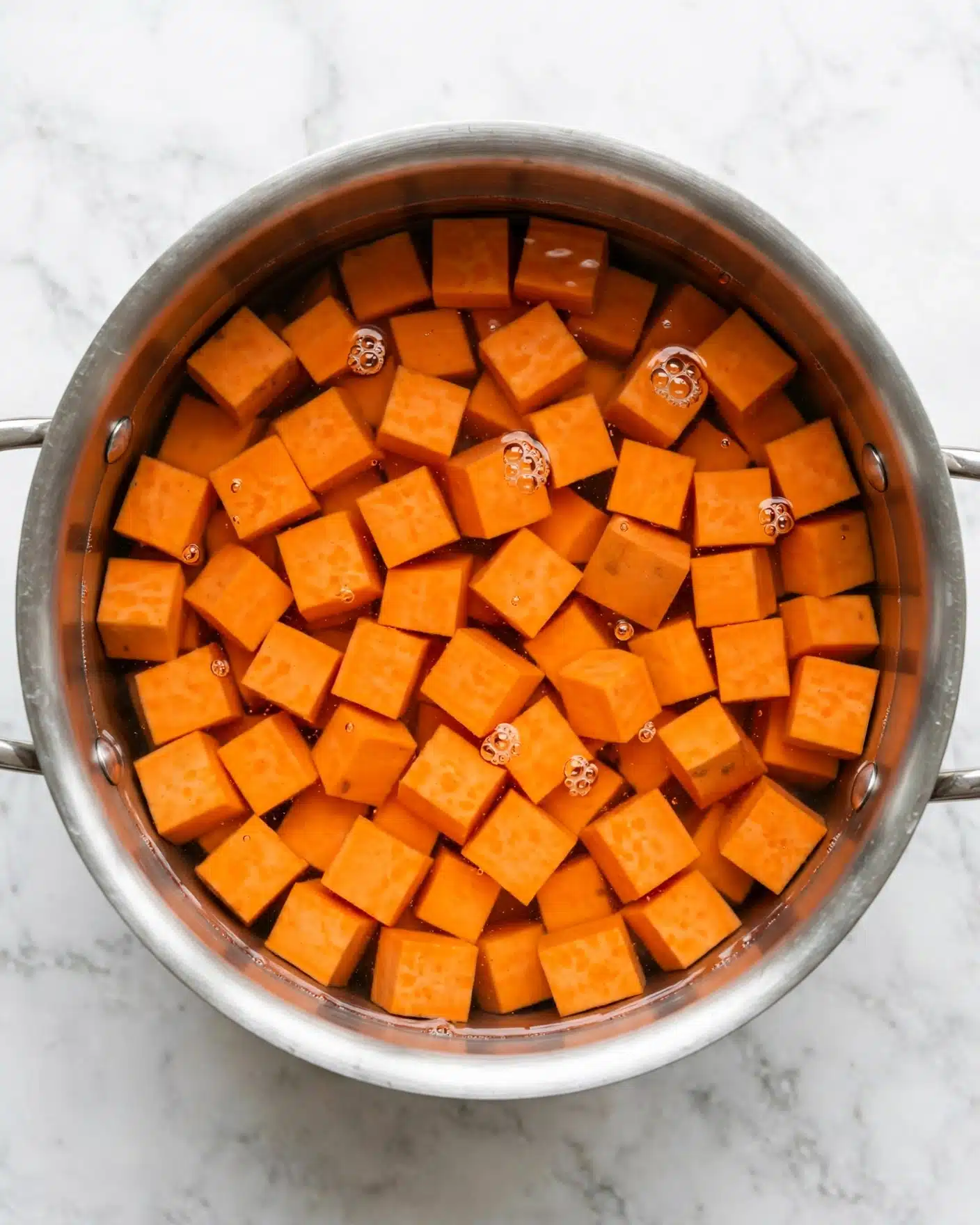 Sweet Potato Cornbread Recipe 4 A silver metal pot shown from above holds bright orange sweet potato cubes submerged in clear water, with a few small bubbles floating on the surface. The sweet potato pieces are roughly cube-shaped and fill the pot almost to the top, with the water covering them completely. The pot rests on a clean white marbled surface. photo taken with an iphone --ar 4:5 --v 7
