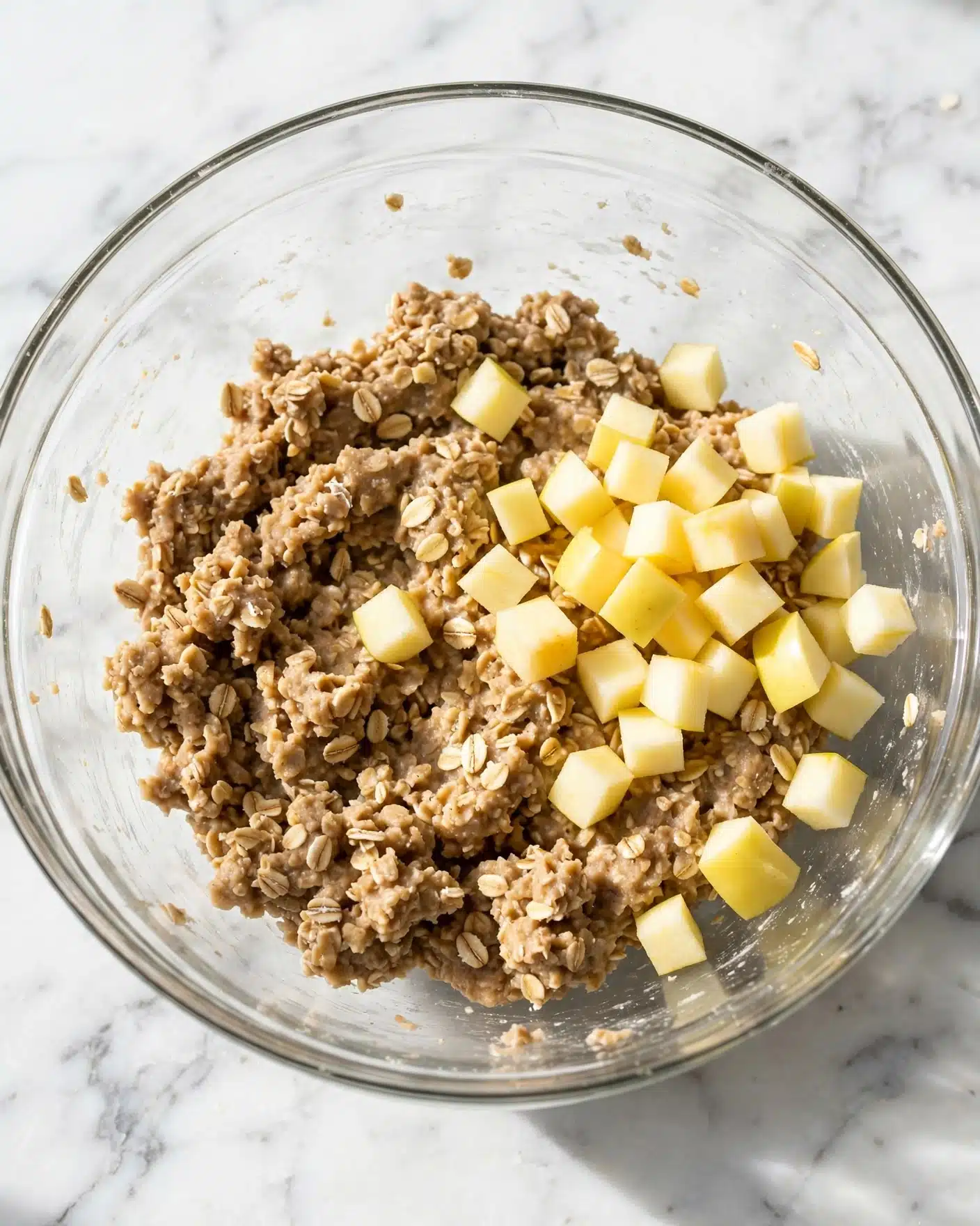 Apple Cinnamon Oatmeal Cookies Recipe 4 In a clear glass bowl, there is a mixture of oatmeal-like dough at the bottom, light brown in color and textured with small oat pieces. On top of this mixture, there are small cubes of pale yellow apple pieces, scattered over the dough mostly on one side. The bowl sits on a white marbled surface, giving a clean and bright look to the setting. The image is clear and bright with soft natural light. photo taken with an iphone --ar 4:5 --v 7