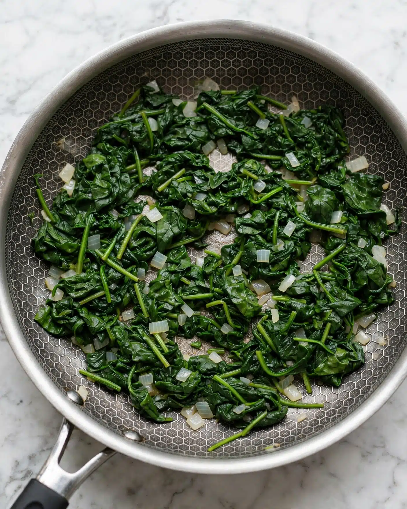 Vegetarian Pumpkin Spinach Lasagna Recipe 4 The image shows a silver frying pan with a hexagonal pattern inside, filled with cooked dark green spinach leaves mixed with small pieces of translucent white onions. The spinach leaves are soft and slightly wilted, covering most of the pan's surface evenly with some visible stems. The pan rests on a white marbled surface, with a small part of the handle visible at the bottom left. photo taken with an iphone --ar 4:5 --v 7