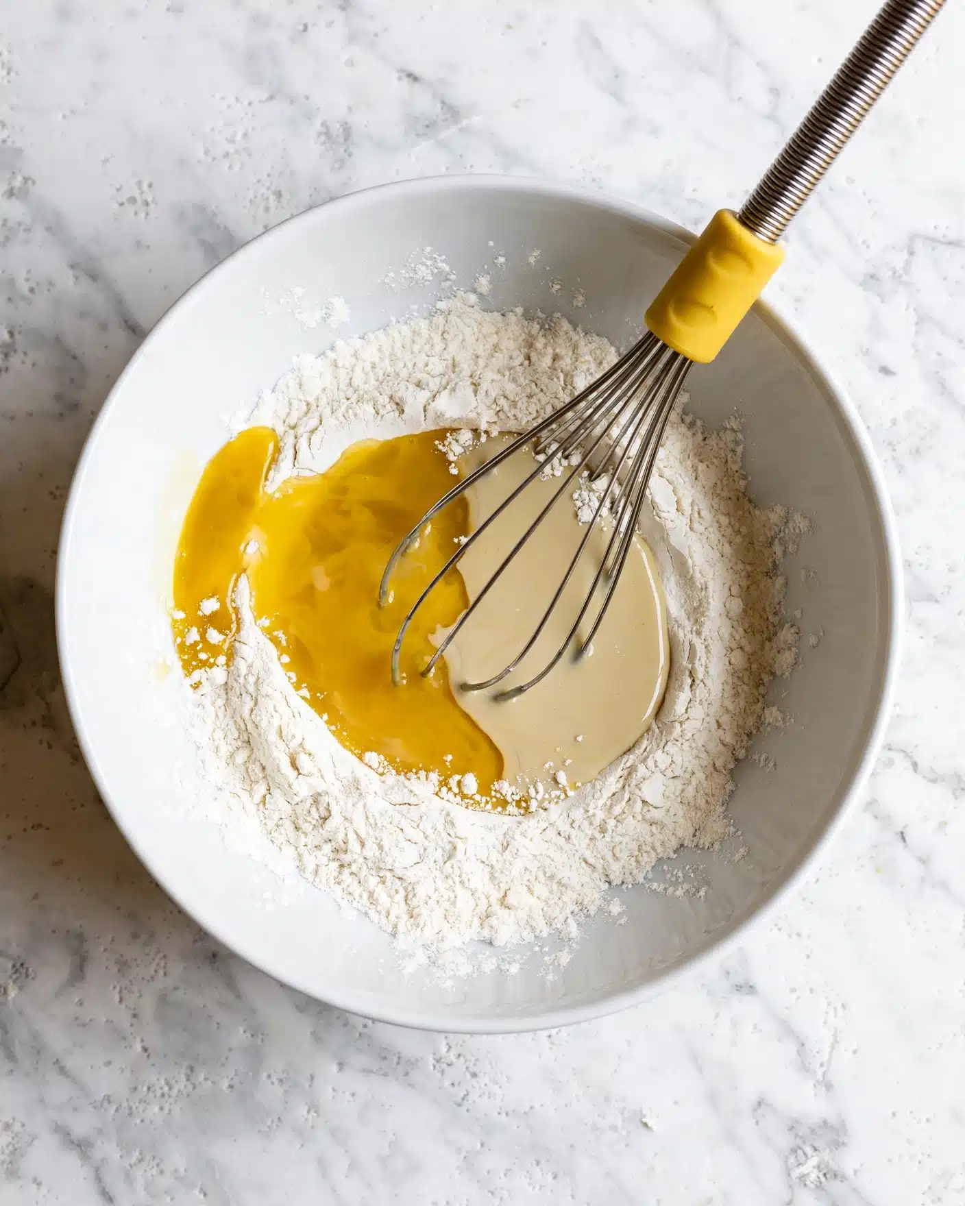 A white bowl on a white marbled surface contains a mix of dry flour and wet ingredients. The mixture shows one main layer of white flour with two pools of wet liquid ingredients next to each other, one golden yellow and the other pale beige. A metal whisk with a yellow handle is placed inside the bowl, touching the ingredients. The photo is taken from above. photo taken with an iphone --ar 4:5 --v 7