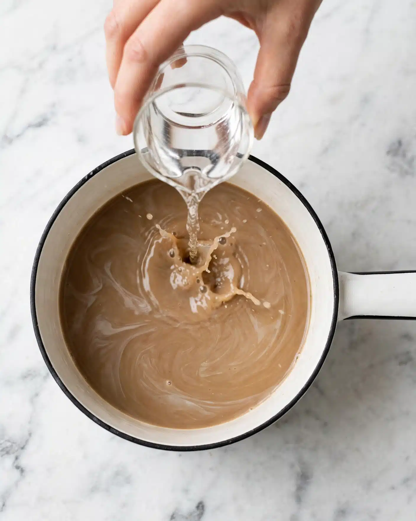 A white pot with a black rim is shown from above, filled with a smooth, light brown liquid that has swirls of a slightly lighter shade on the surface. A woman's hand is pouring a clear liquid from a small glass container into the pot, creating a small splash where the two liquids meet. The pot is on a white marbled surface, and the handle of the pot extends to the right side of the image. The scene is clean and simple, focusing on the mixture in the pot and the pouring action. photo taken with an iphone --ar 4:5 --v 7