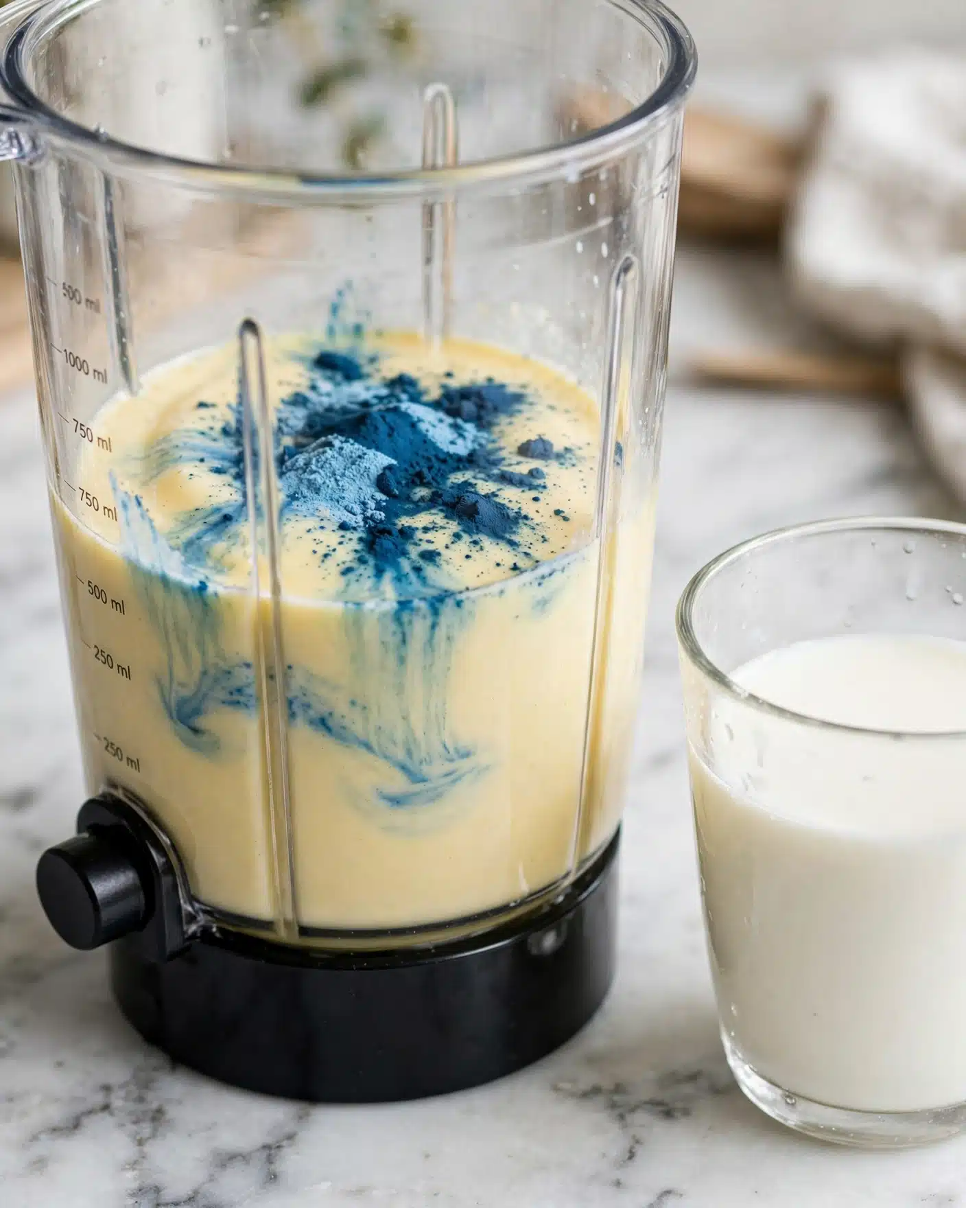 Blue Milk (Star Wars Inspired!) Recipe 4 A clear blender jar shows a creamy light yellow mixture with a bright blue powder sprinkled on top before blending. The jar sits on a white marbled surface next to a glass cup filled with white liquid. The blue powder is finely textured and partially dissolving into the creamy base. The blender is clear with a black button visible on the left side. photo taken with an iphone --ar 4:5 --v 7